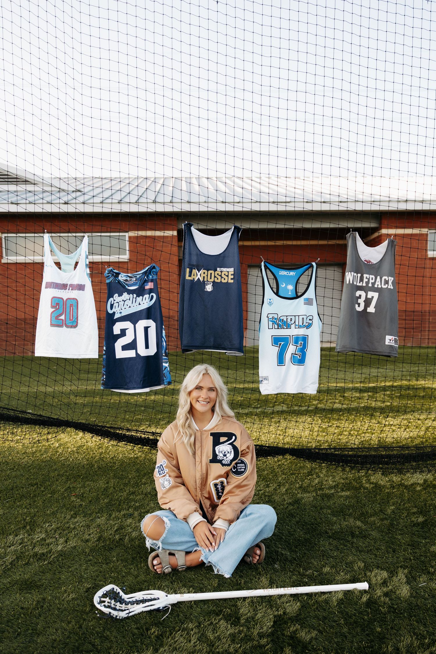 Blonde female lacrosse player sits on turf field holding stick, with five sports jerseys displayed on net behind her.