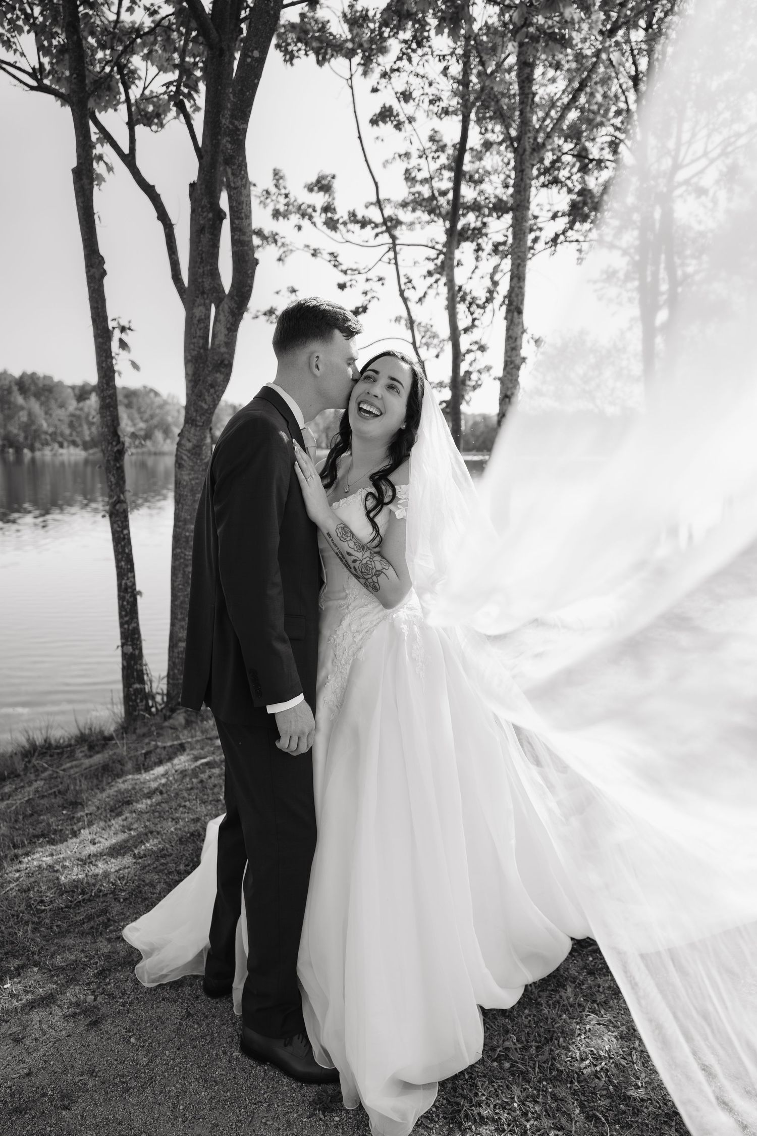 Laughing bride's veil blows in wind as groom kisses her cheek near lakeside trees in black and white.