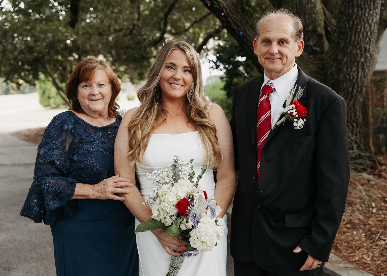 Bride in a white strapless gown holding a red and white bouquet posing with her parents outdoors.
