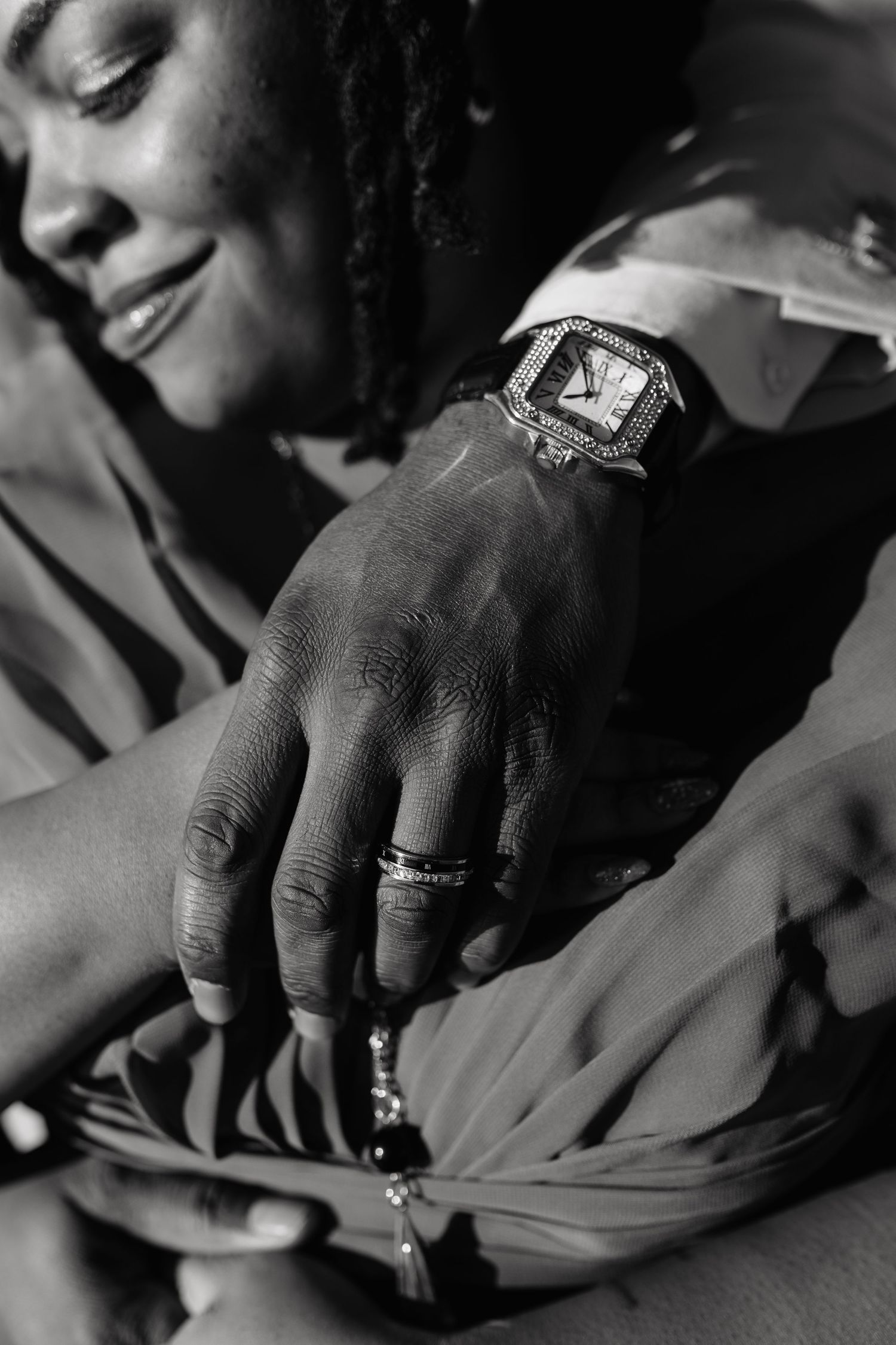 Black and white photo of a man's hand wearing a diamond-encrusted watch and rings resting on a woman's lap.