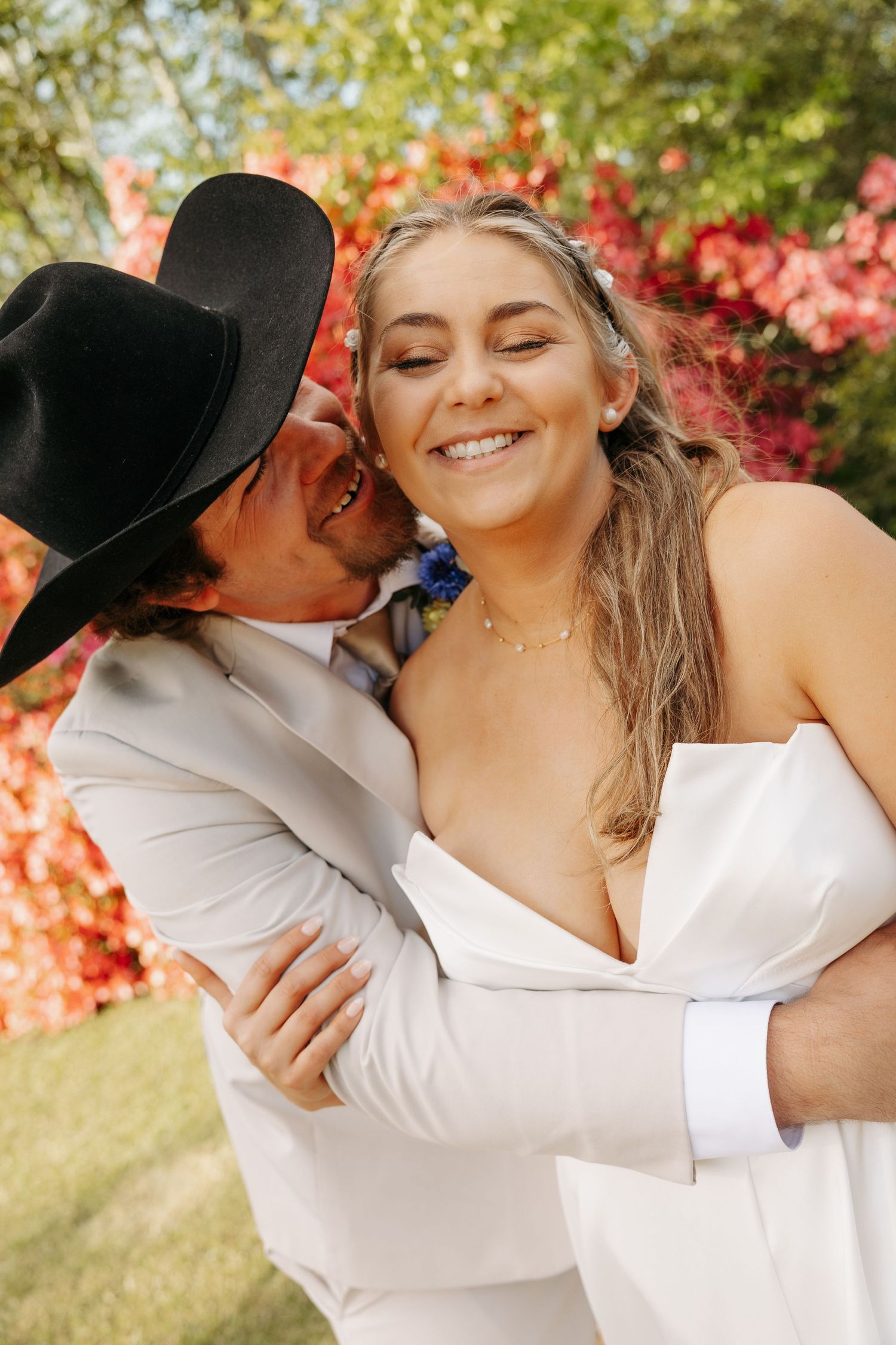 Groom in black cowboy hat kissing smiling bride on cheek in front of vibrant red floral backdrop.
