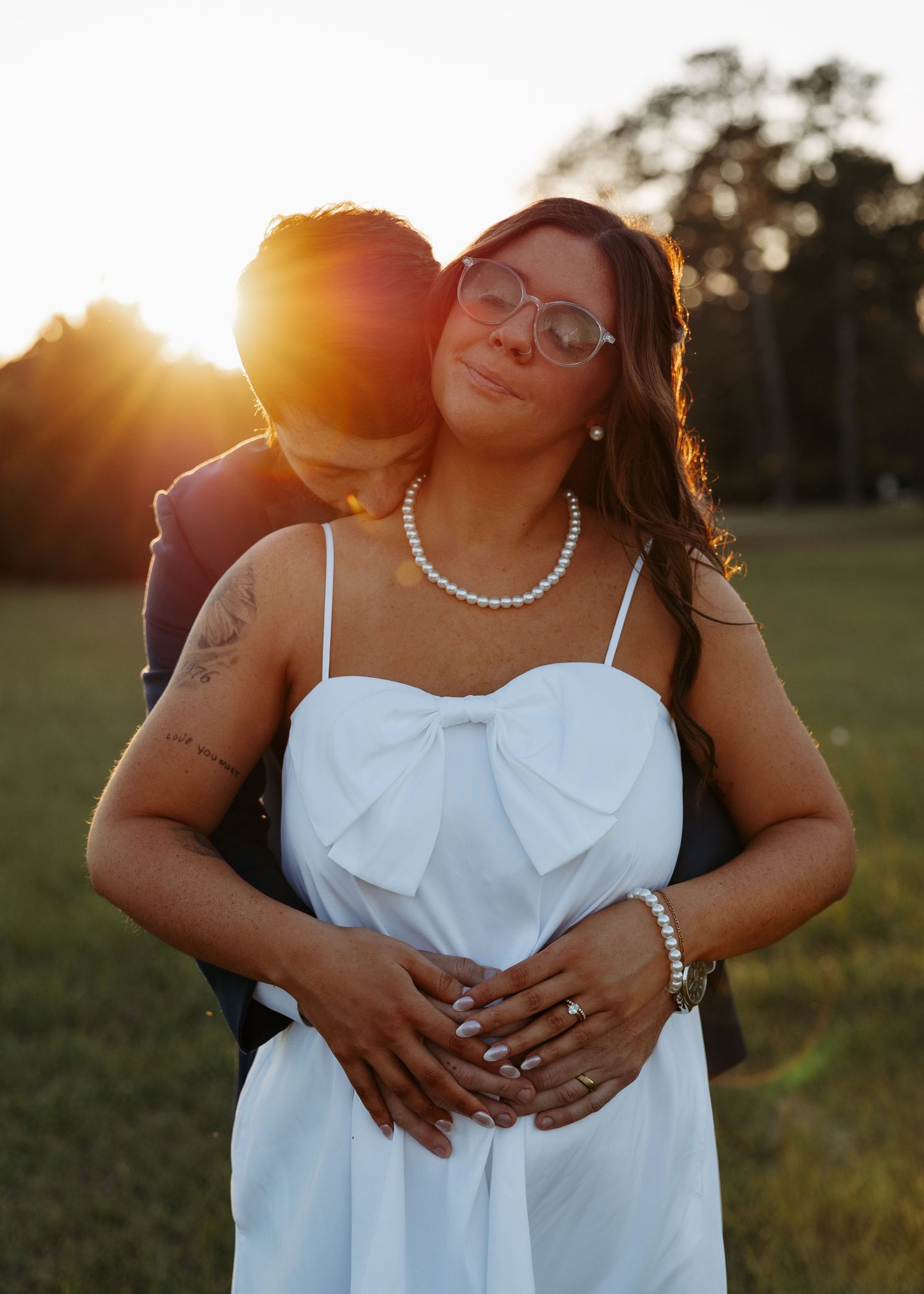 Groom embraces bride from behind at golden sunset during romantic outdoor wedding portraits.