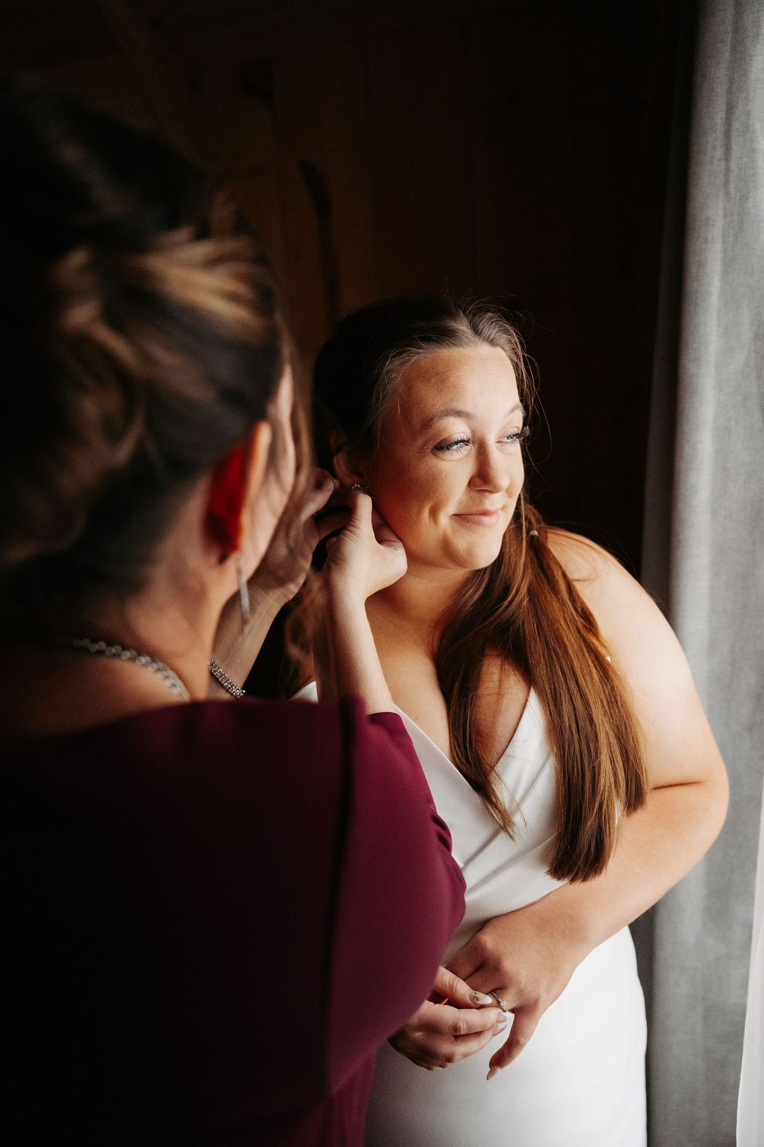 Bride in white gown having earring put on by bridesmaid near window.