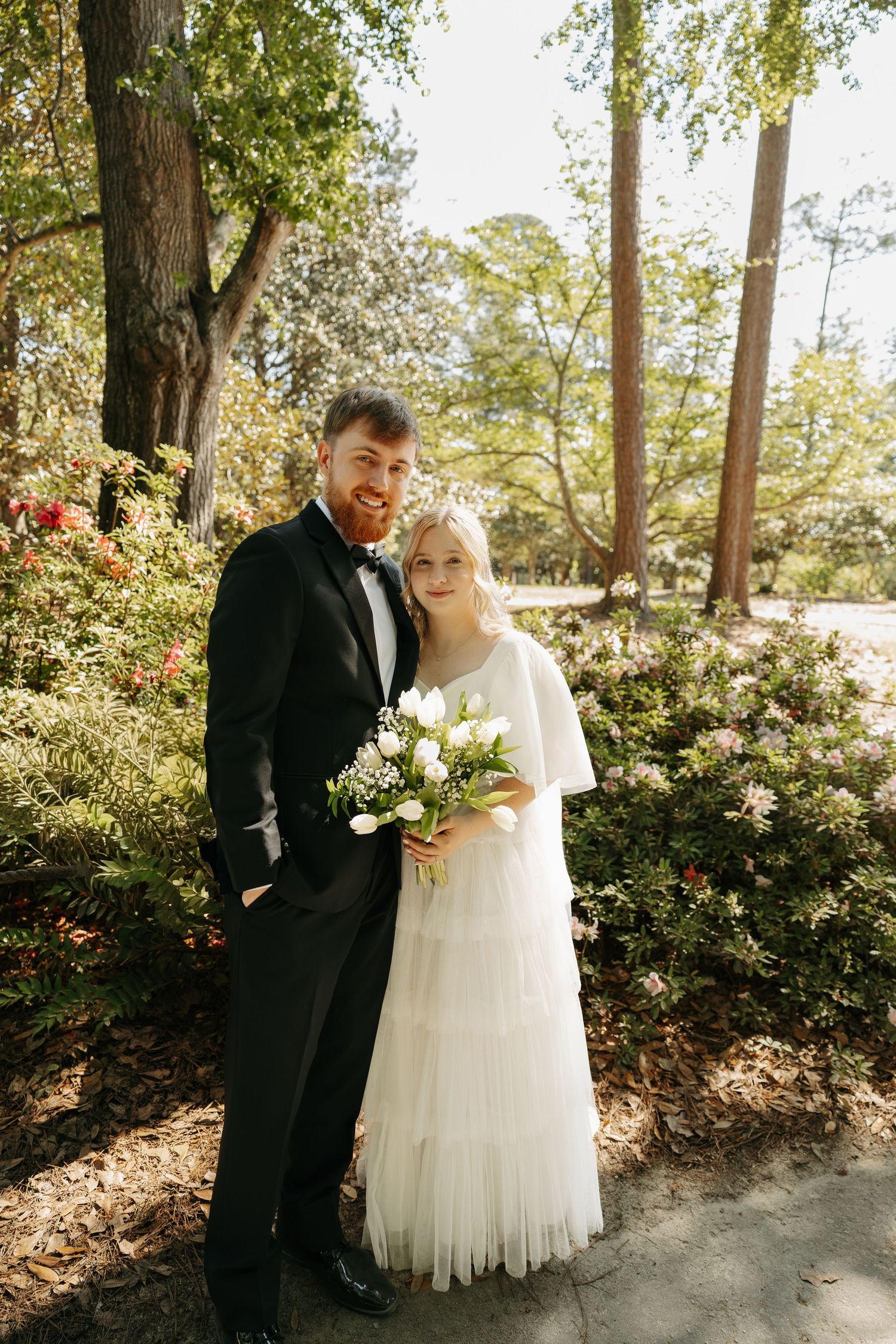 Smiling bride and groom pose together in a sunlit garden, bride holding a white tulip bouquet.