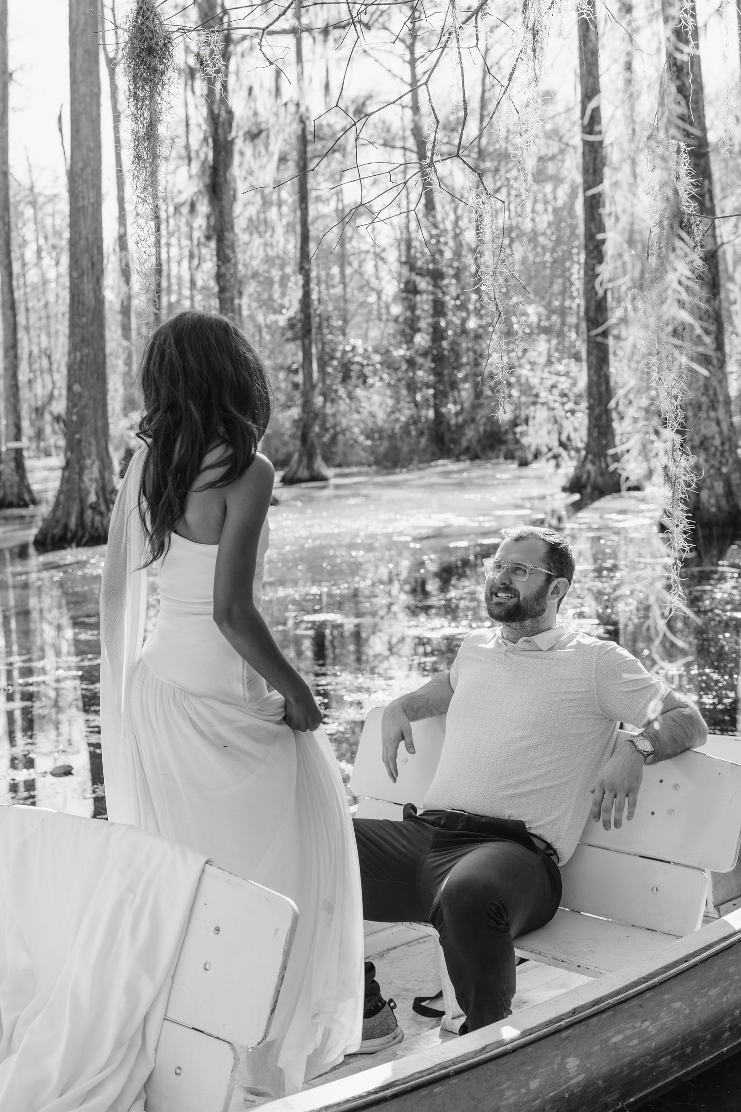 Black and white photo of a couple on a boat in a swamp, woman in white dress standing while man sits looking up at her.