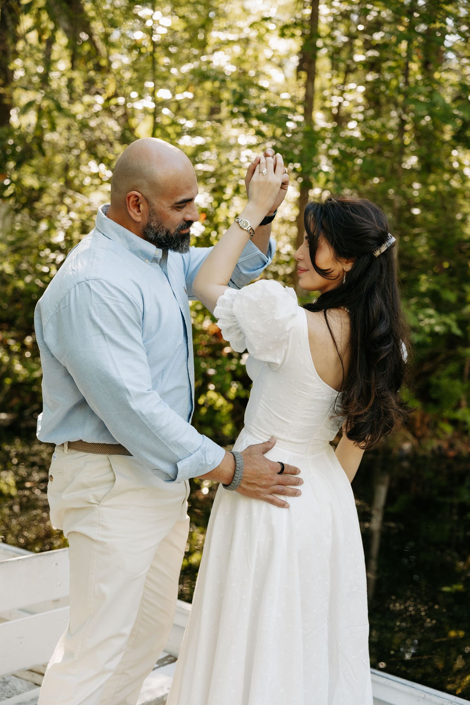 Couple dancing outdoors in forest setting, groom in blue shirt and bride in white dress, sharing romantic moment.