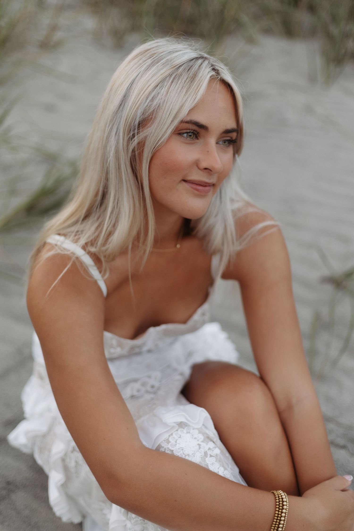 Woman in white lace dress poses on sandy beach with platinum blonde hair and gold bracelet.