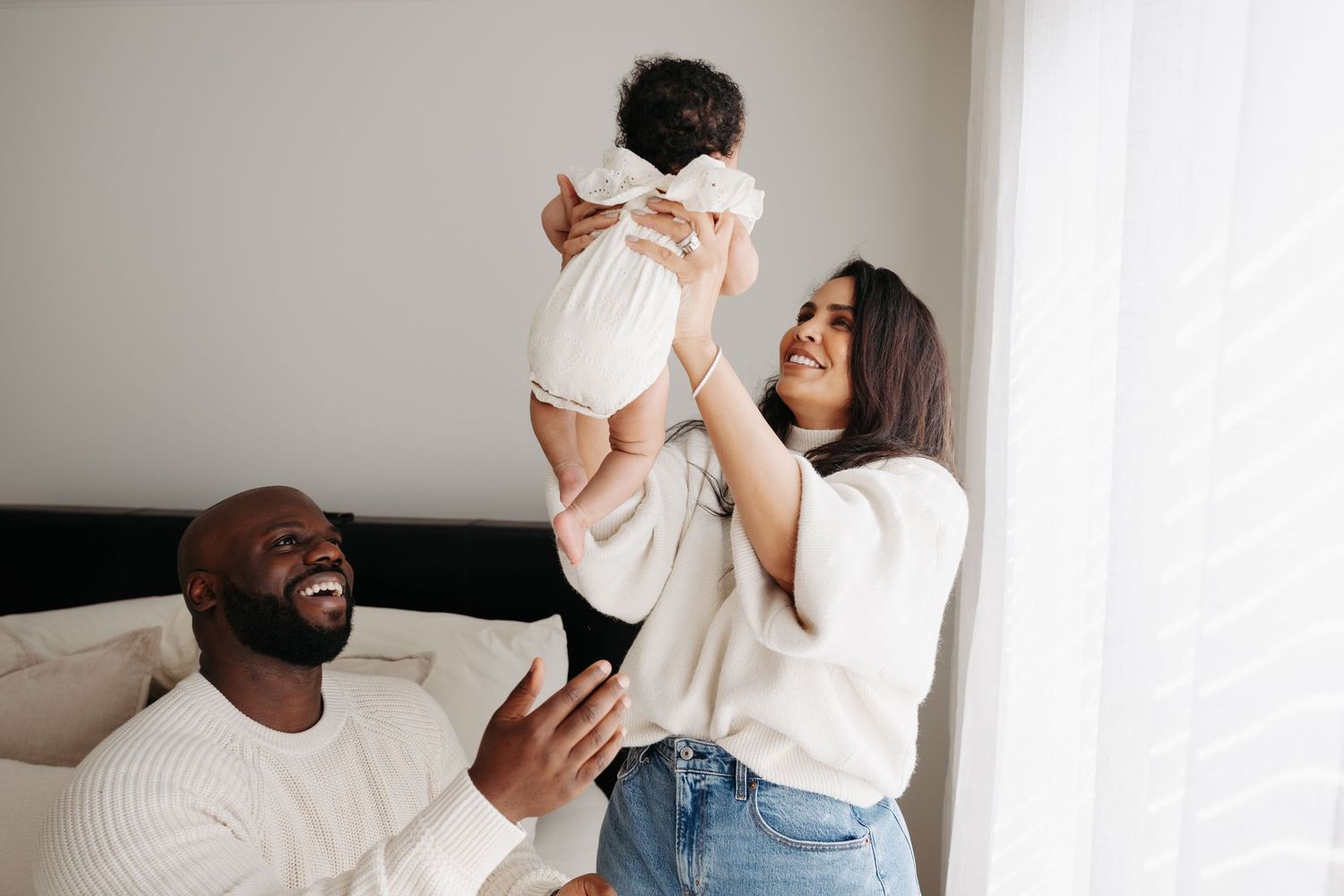 Happy family with parents lifting smiling baby in bright bedroom, sharing joyful moment together.