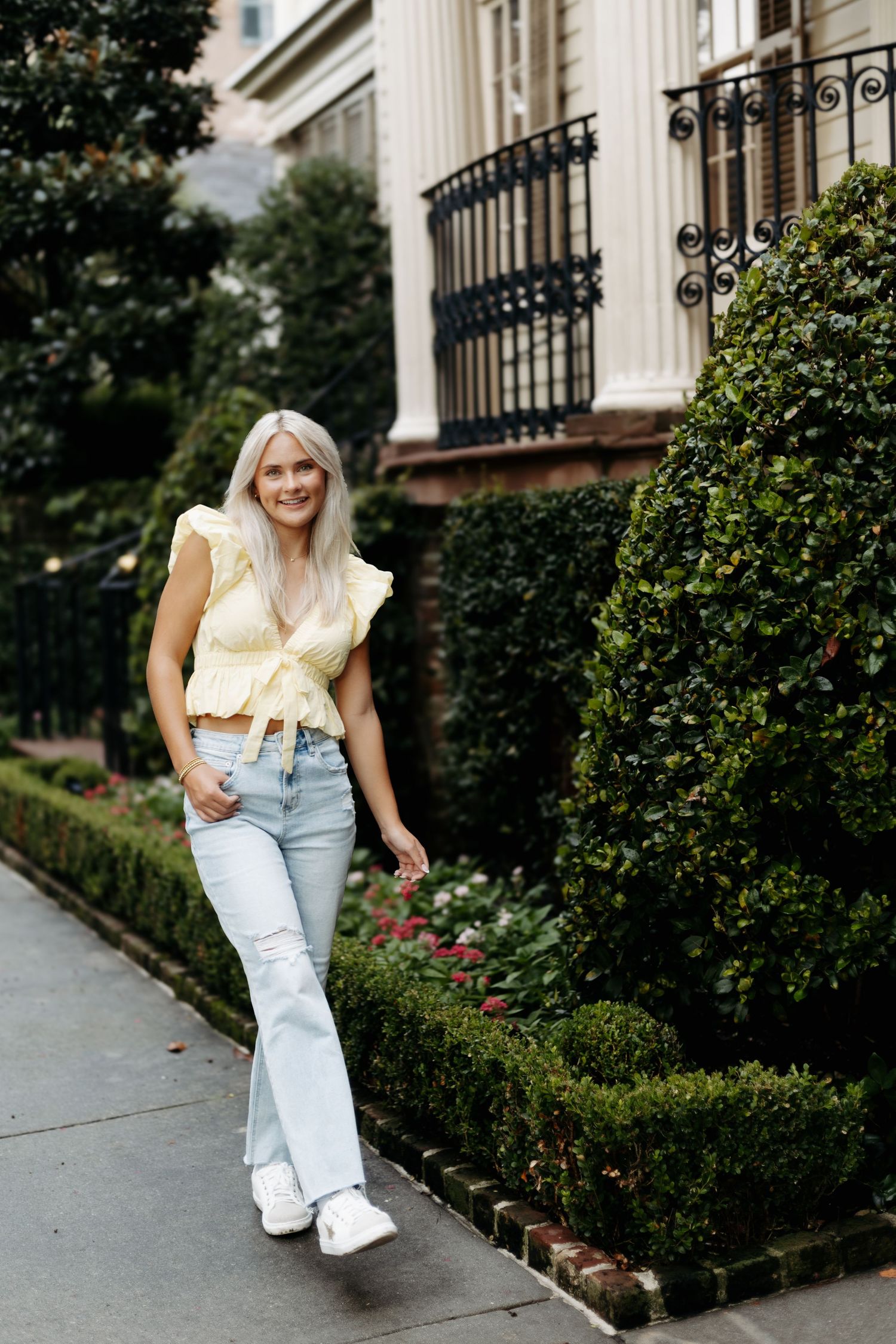 Woman in yellow crop top and light blue flare jeans smiling outdoors near lush green hedges and elegant building.