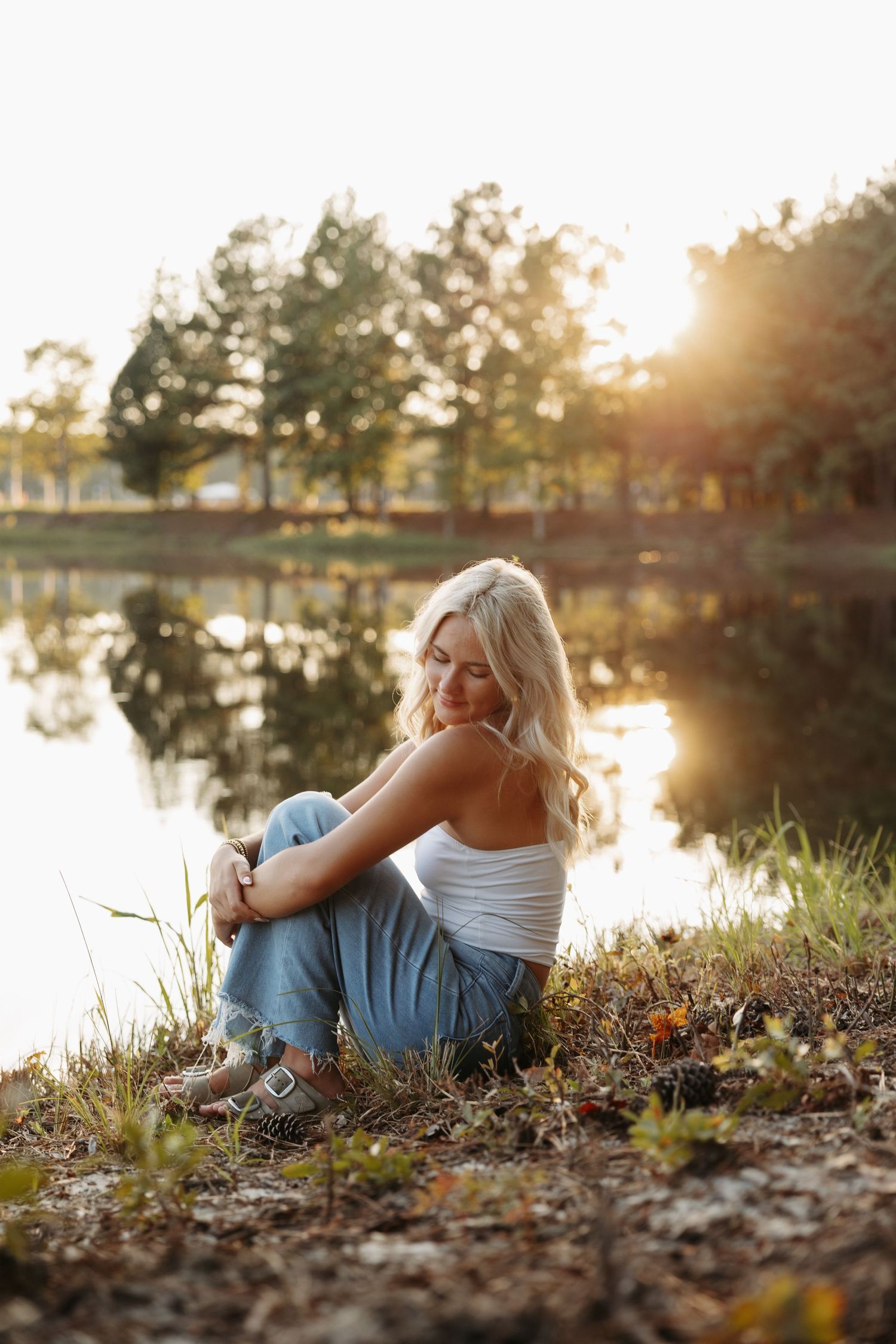 Blonde woman sitting by a reflective lake at golden hour, wearing white top and jeans, surrounded by lush green trees.