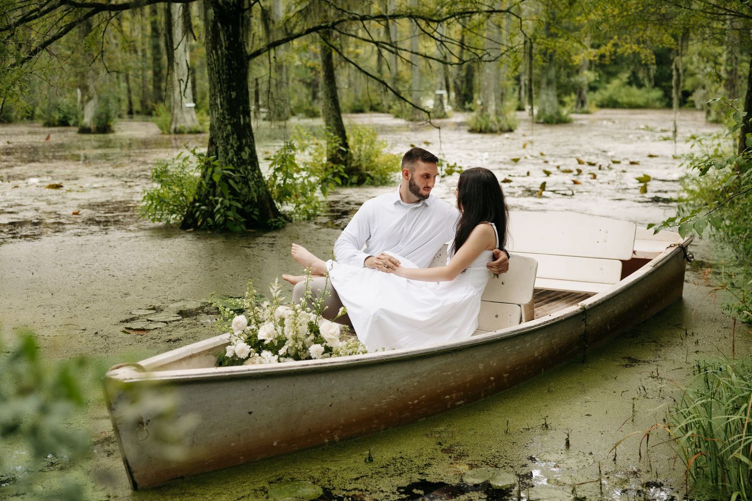 Romantic couple sitting in a wooden canoe surrounded by lush green trees and lily pads on a serene swamp.