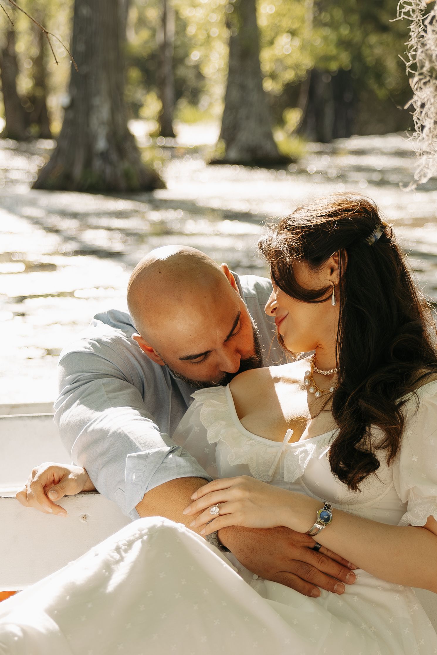 A bald groom kisses his bride's forehead as they share a tender moment outdoors in dappled sunlight.