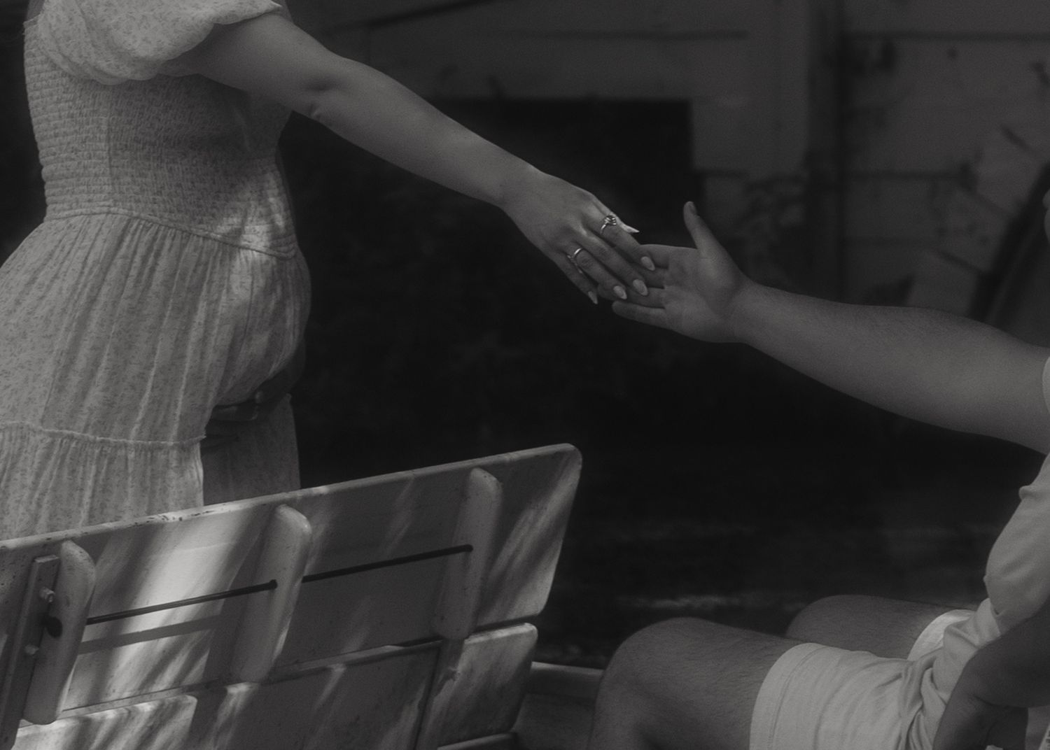 Black and white photo of two people reaching out to hold hands near a chair outdoors.