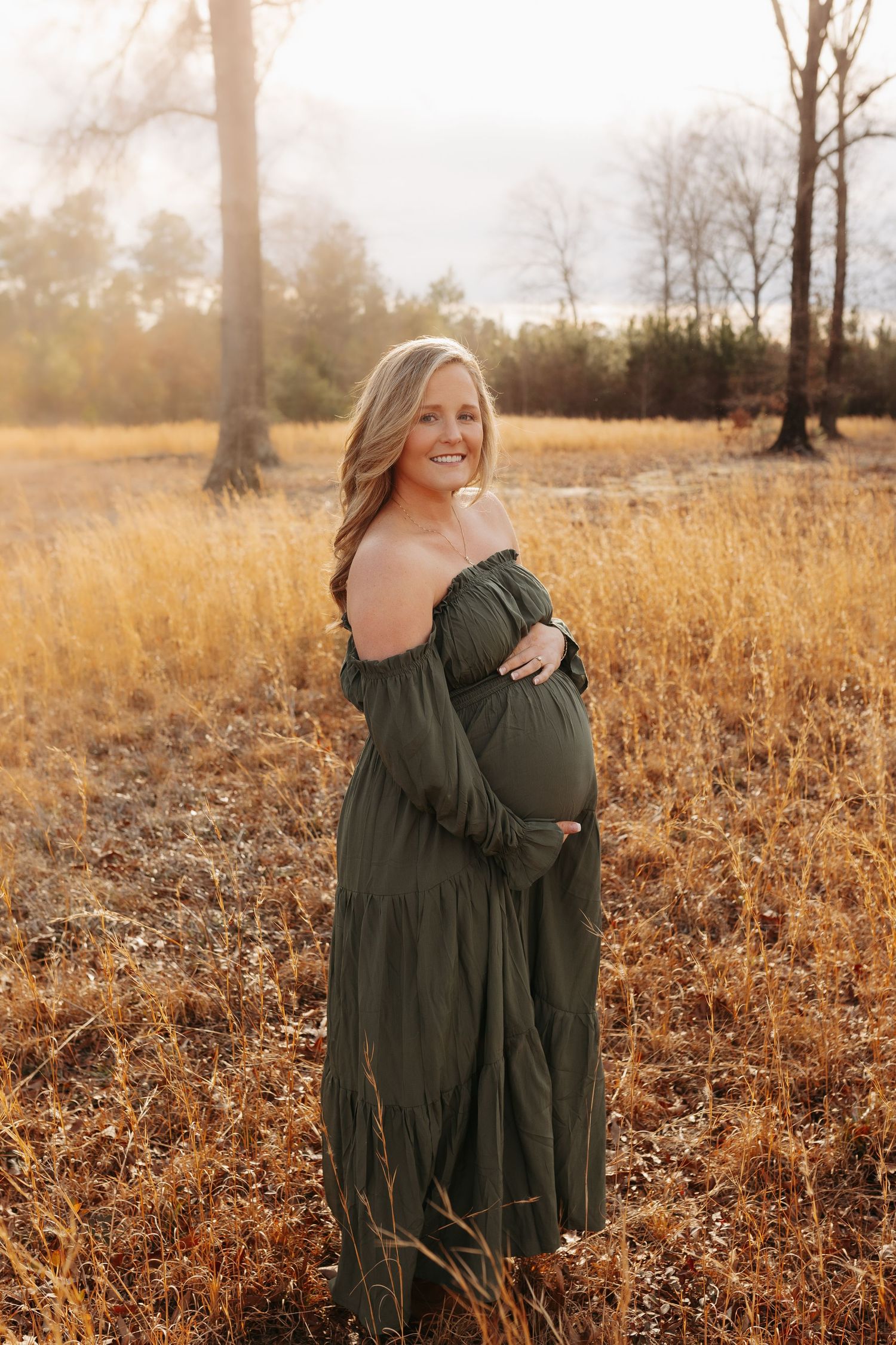A person in a dark green dress poses in a golden autumn field at sunset with warm glowing light.