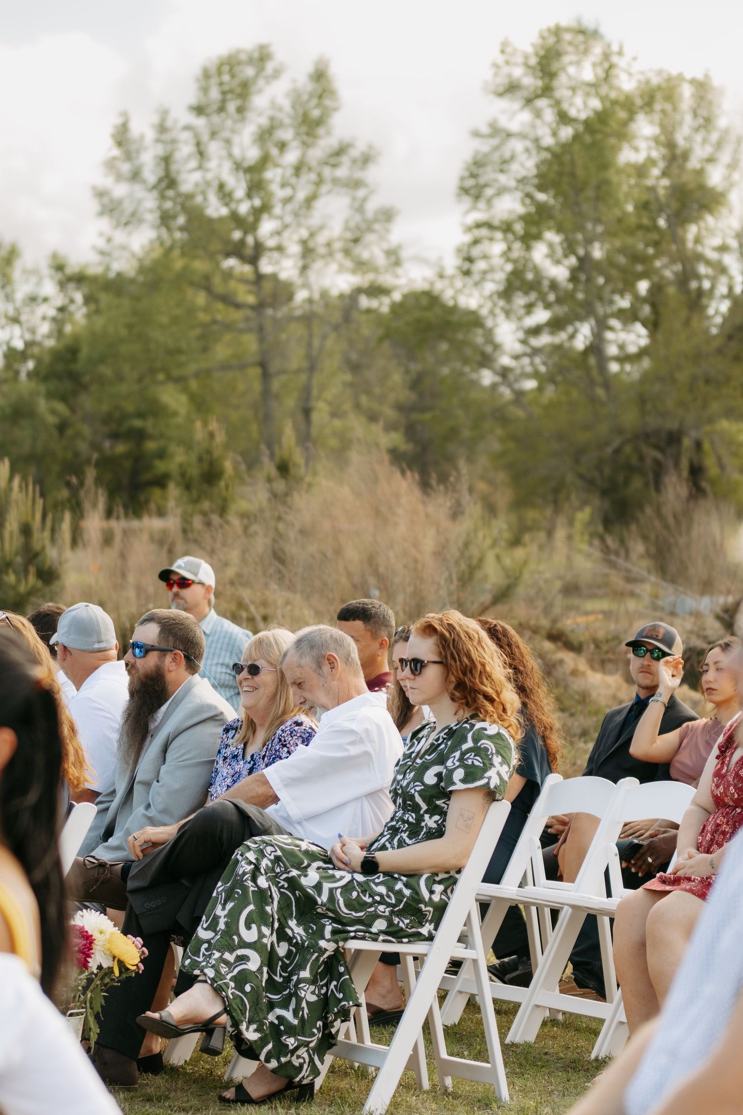 Guests seated in white chairs at an outdoor wedding ceremony surrounded by trees and natural landscape.