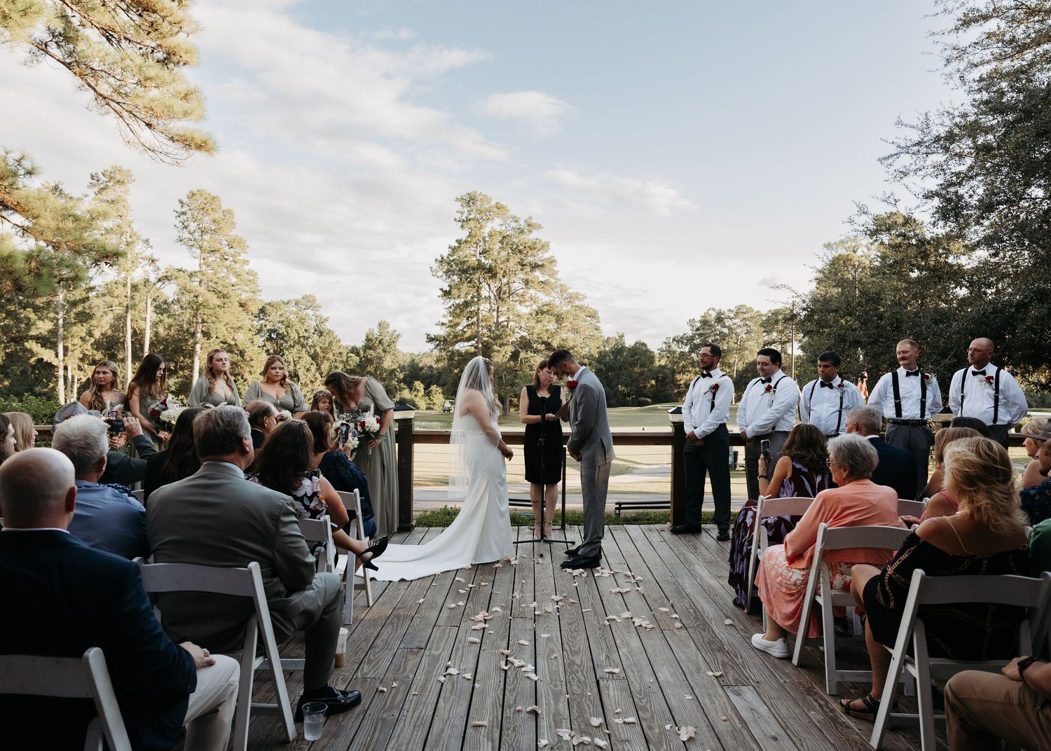 Outdoor wedding ceremony on wooden deck with guests seated amid lush trees.