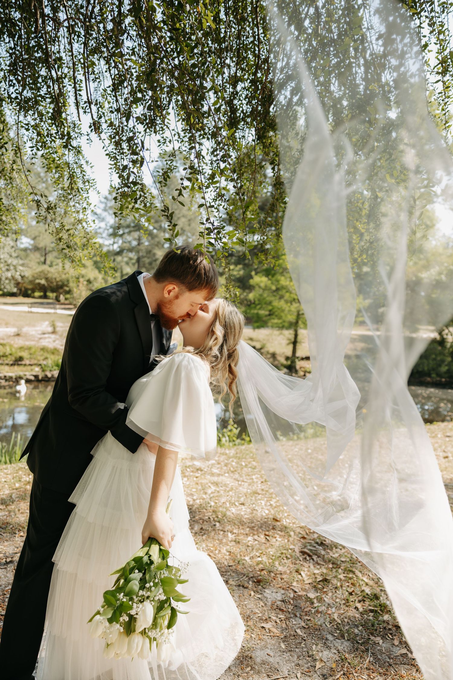 Bride and groom sharing a romantic kiss under a weeping willow tree, with veil flowing in the breeze.