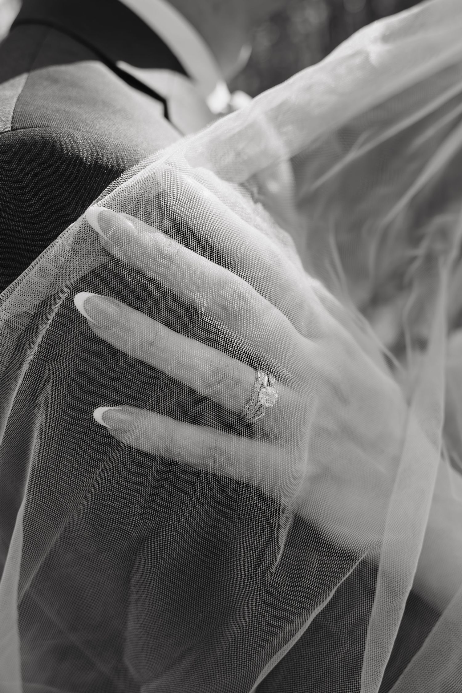 Black and white close-up of a bride's hand with diamond wedding ring resting on groom's shoulder beneath a sheer veil.
