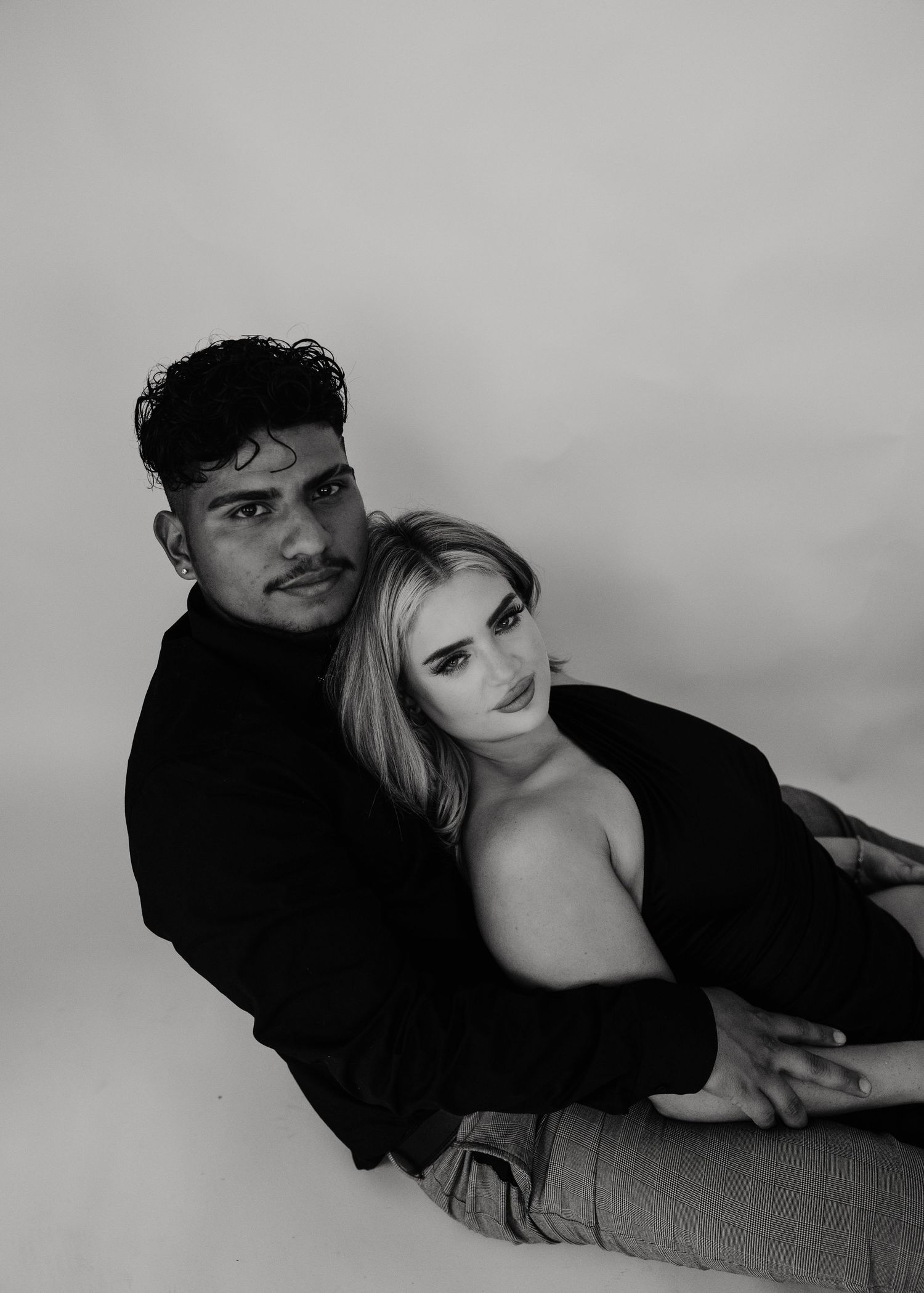 Black and white studio portrait of a couple sitting together, man in black sweater embracing woman with blonde hair.