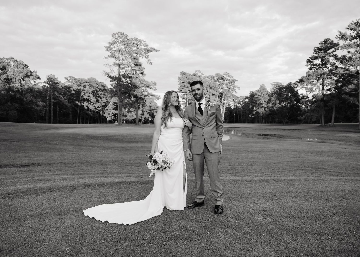 Black and white photo of a bride and groom standing on a golf course, bride holding a bouquet.