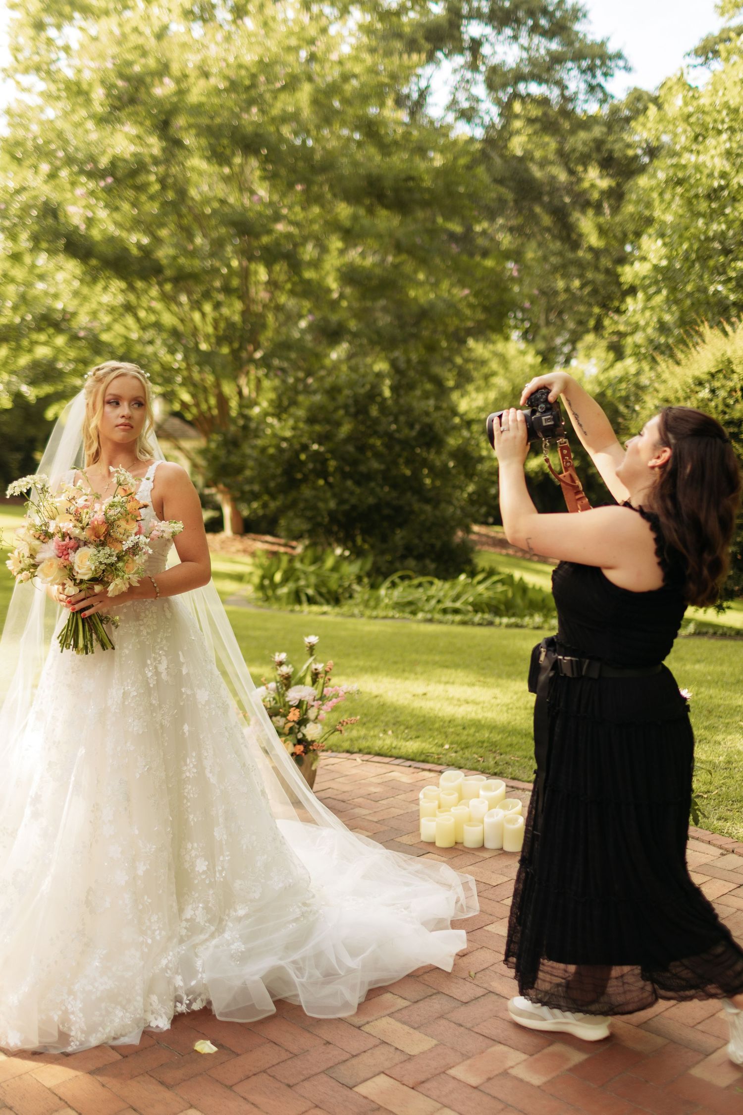 A photographer in black captures a bride in a white lace gown holding a bouquet in a lush garden setting.