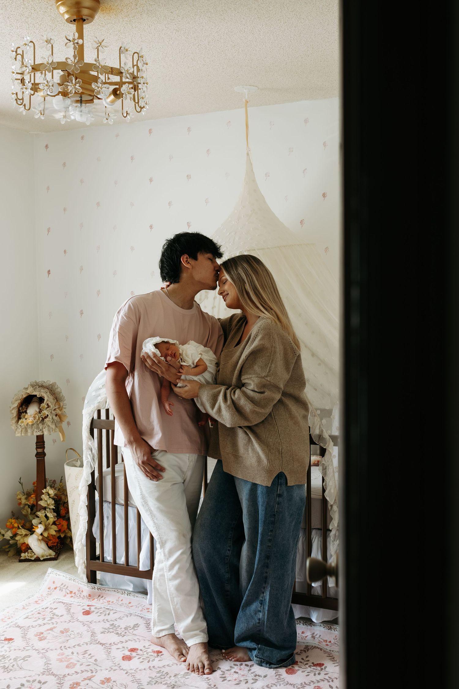 A couple shares a tender moment holding their newborn in a softly decorated nursery with a vintage chandelier.