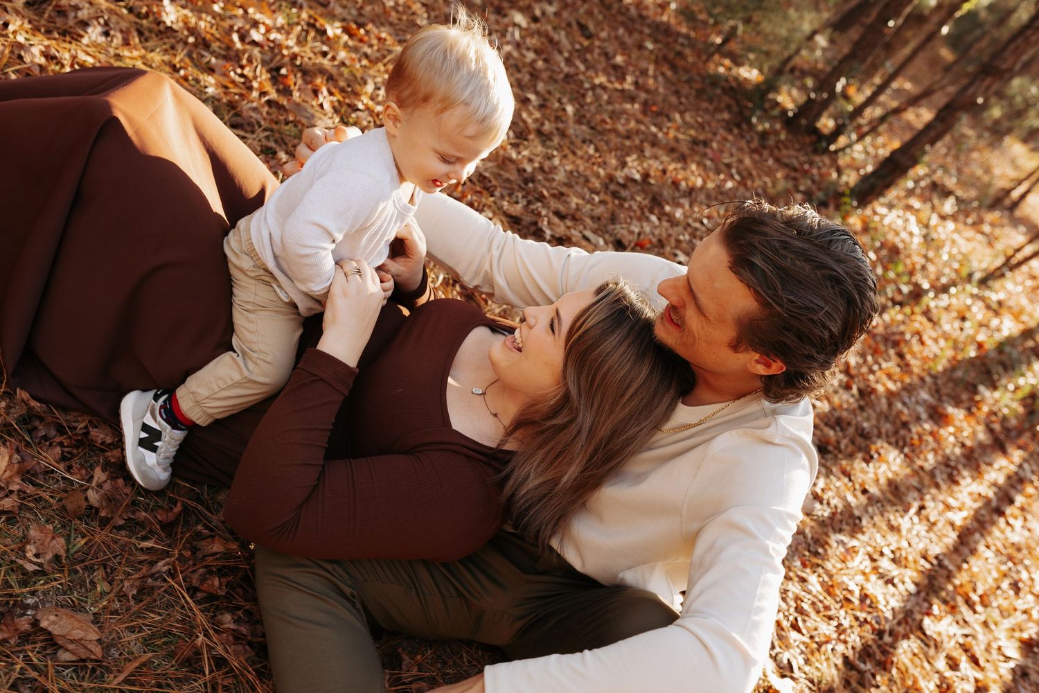 Family enjoying autumn outdoors, mother lying on fallen leaves with baby and father in a warm fall forest setting.