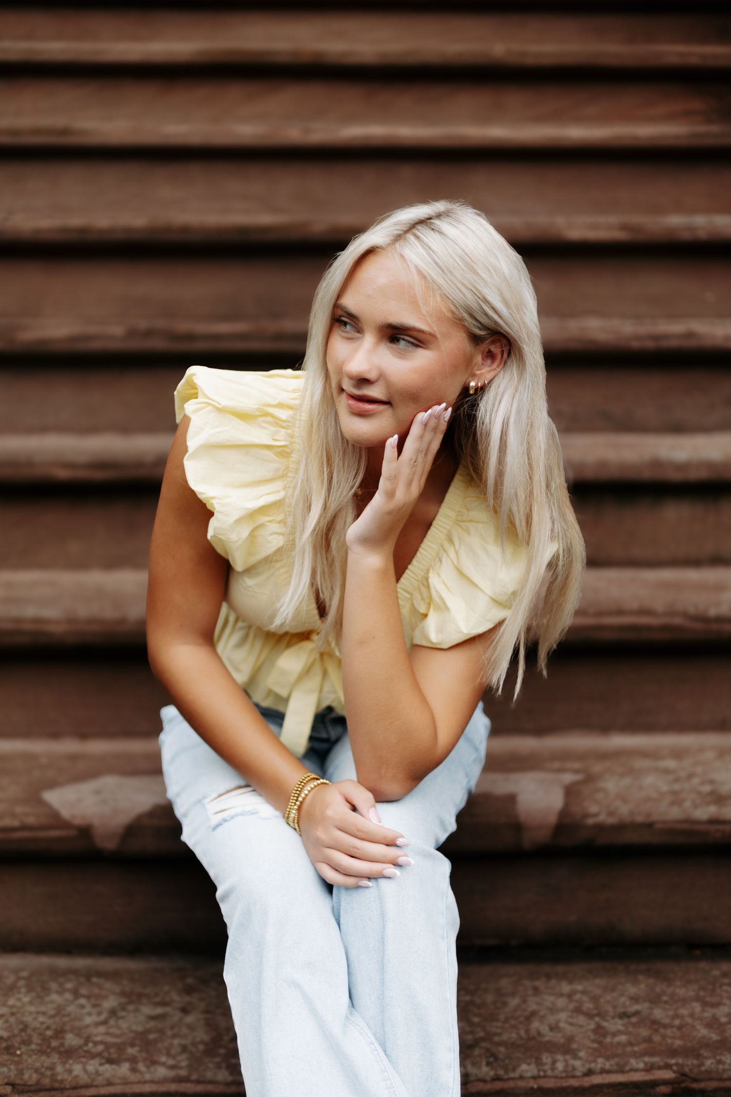 A casual portrait showing someone wearing a yellow ruffled top and white pants against a rustic wood background.