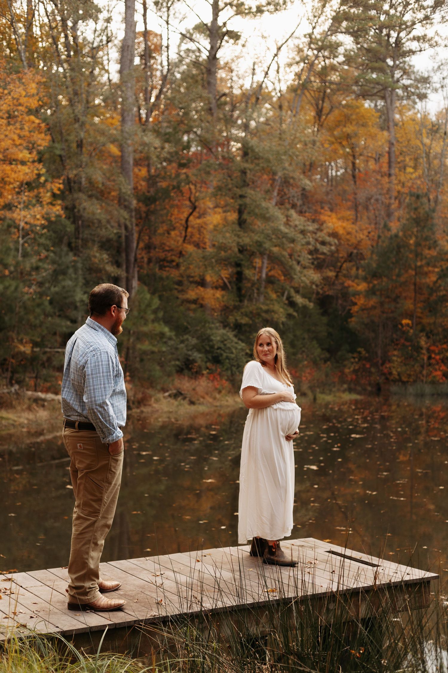 Pregnant woman in white dress stands on autumn lakeside dock with partner among colorful fall foliage.