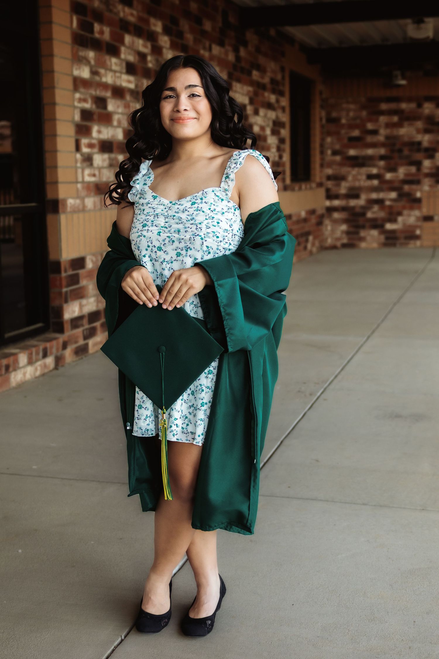 A graduate in floral dress and green graduation gown poses against brick wall.