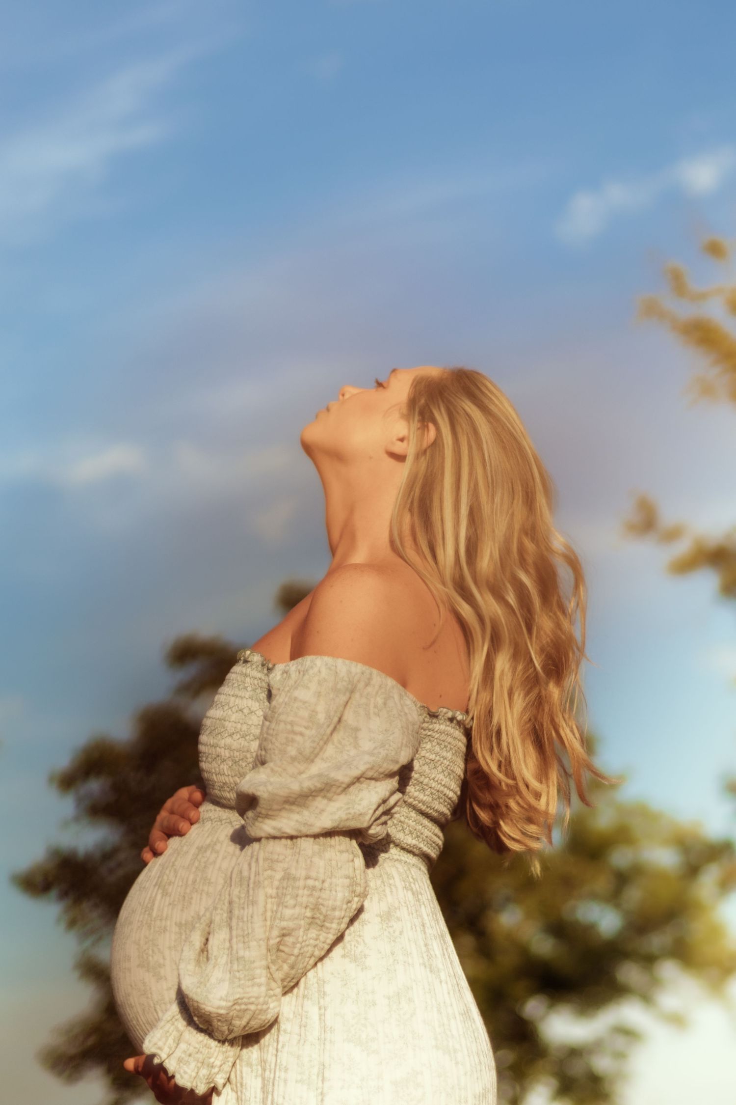 Side profile of pregnant figure in white dress against blue sky and sunset.