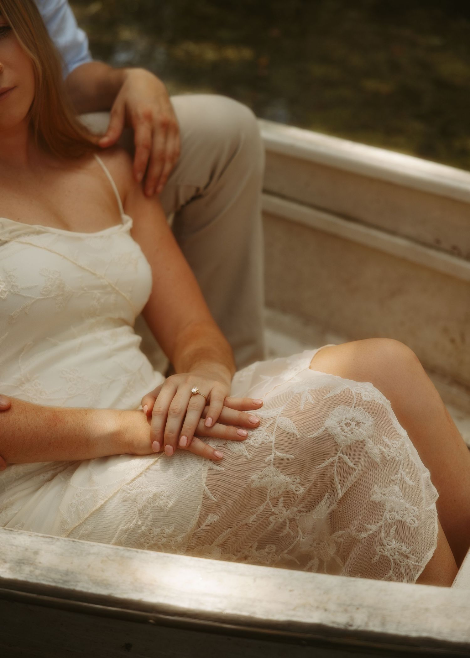 A woman in a white lace dress reclines in a boat, hands intertwined with a partner in a romantic outdoor setting.