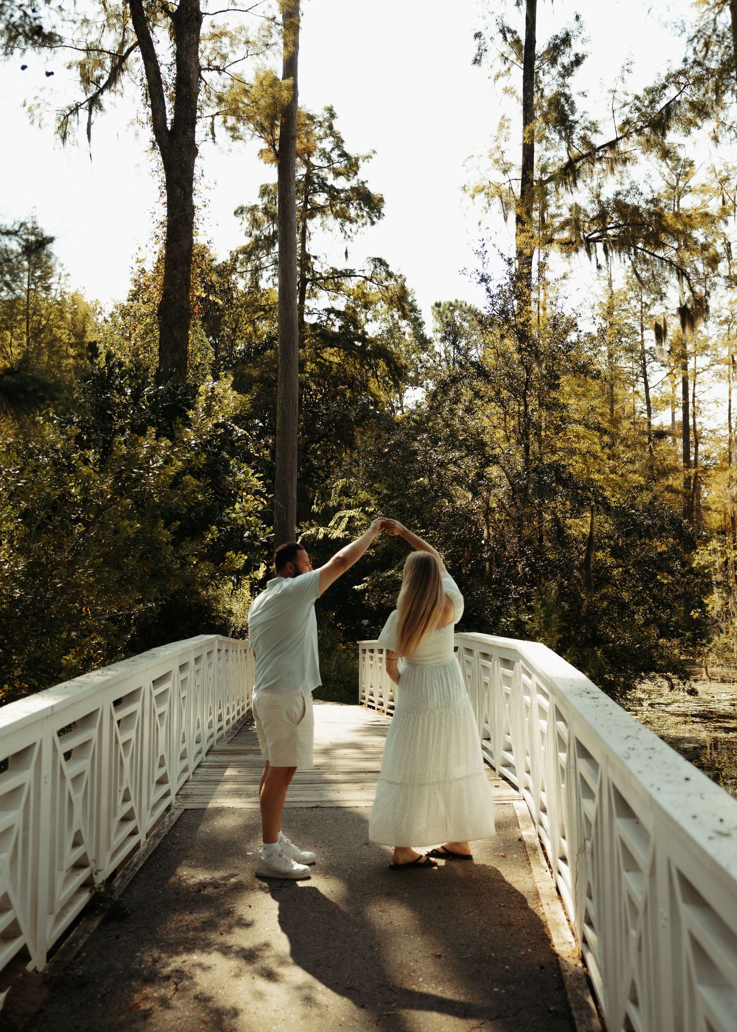 Couple dancing on a white bridge surrounded by lush green trees in a sunlit forest setting.