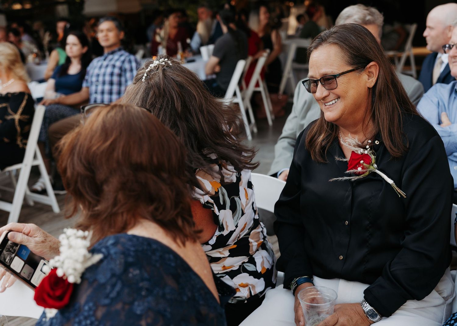 Women in formal attire smiling and conversing at an outdoor event with white chairs.