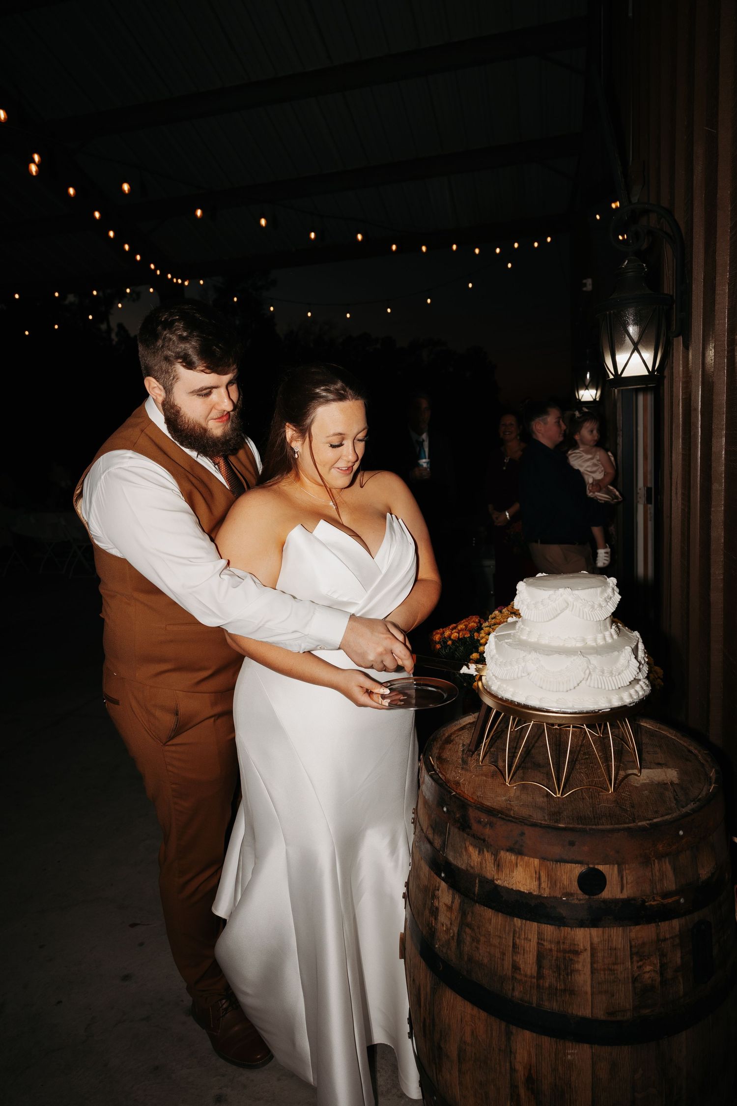 Bride and groom cutting their wedding cake displayed on a rustic barrel under string lights at an outdoor evening reception.