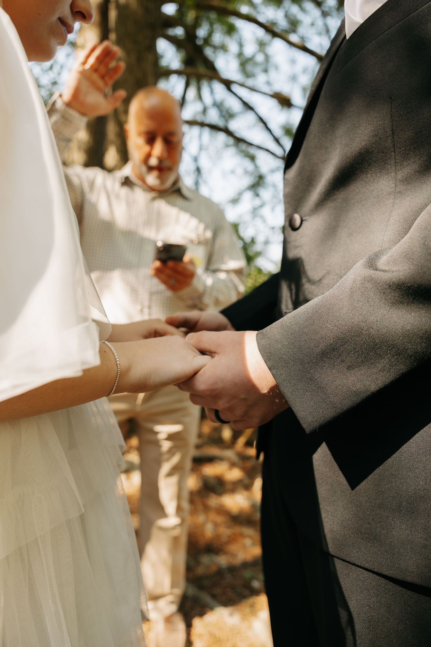 Couple exchanging rings during an outdoor wedding ceremony in a forest, with an officiant reading in the background.