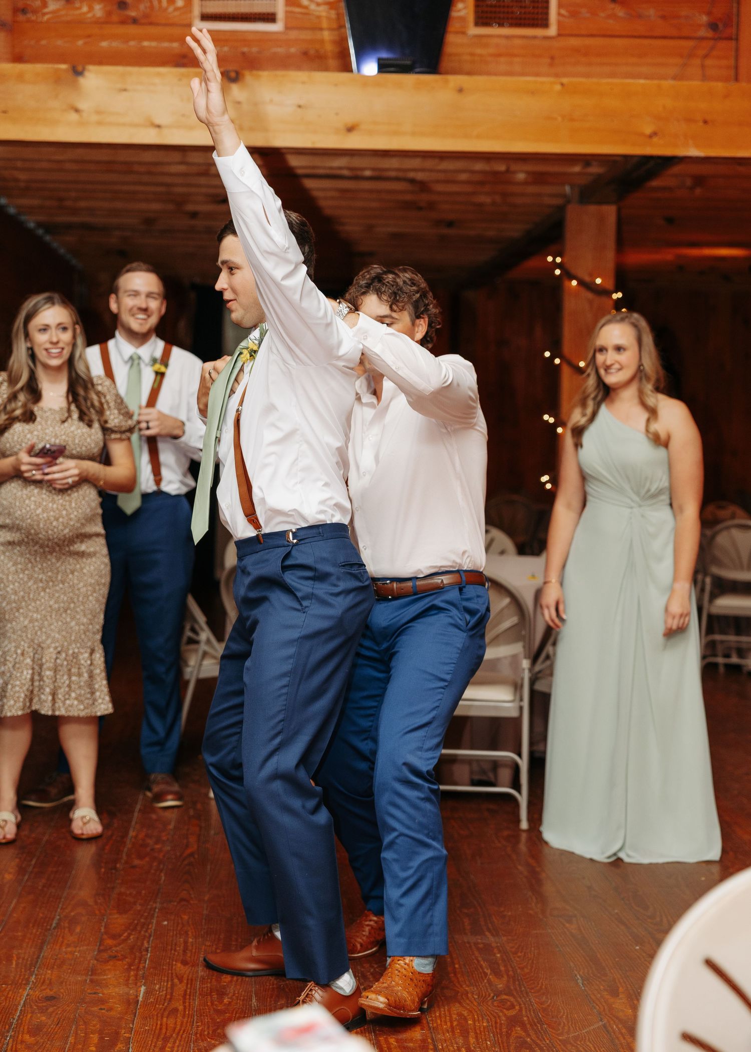 Two groomsmen in blue pants and suspenders dance energetically at a rustic wedding reception while guests smile in background.
