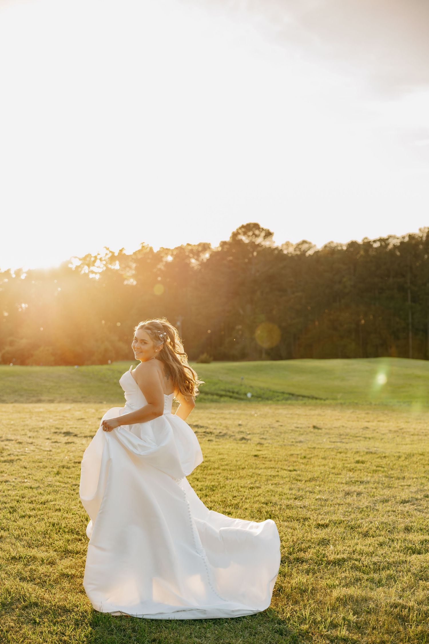 Bride in a flowing white strapless gown twirls in a sunlit open field during golden hour.
