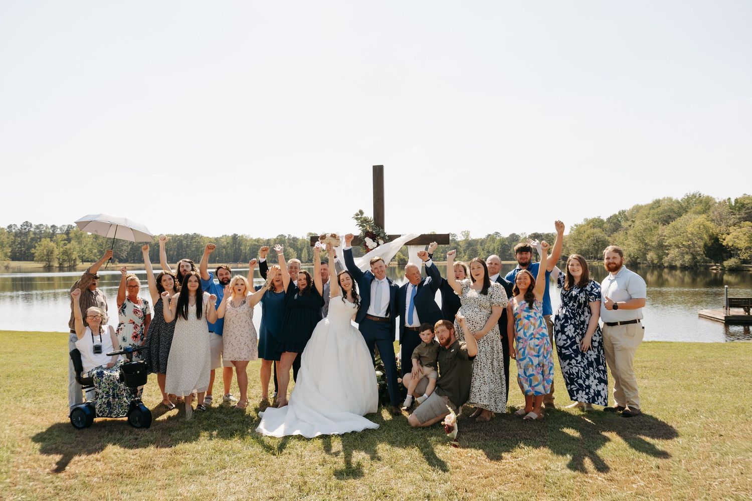 Wedding group photo with bride and groom celebrating outdoors by a lake with a cross backdrop on a sunny day.