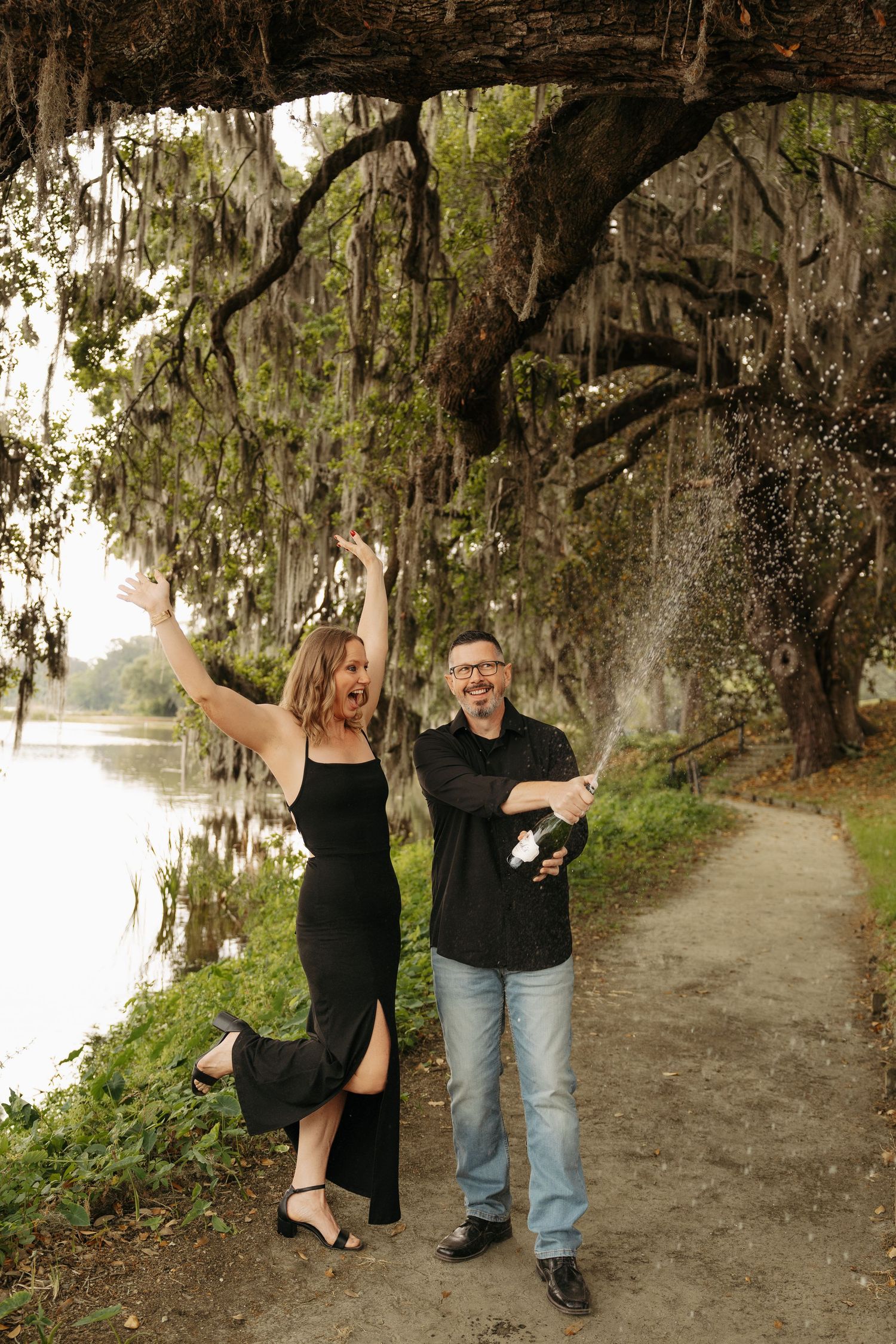 Couple celebrating outdoors under moss-draped oak trees, popping champagne by a river, both dressed in black.