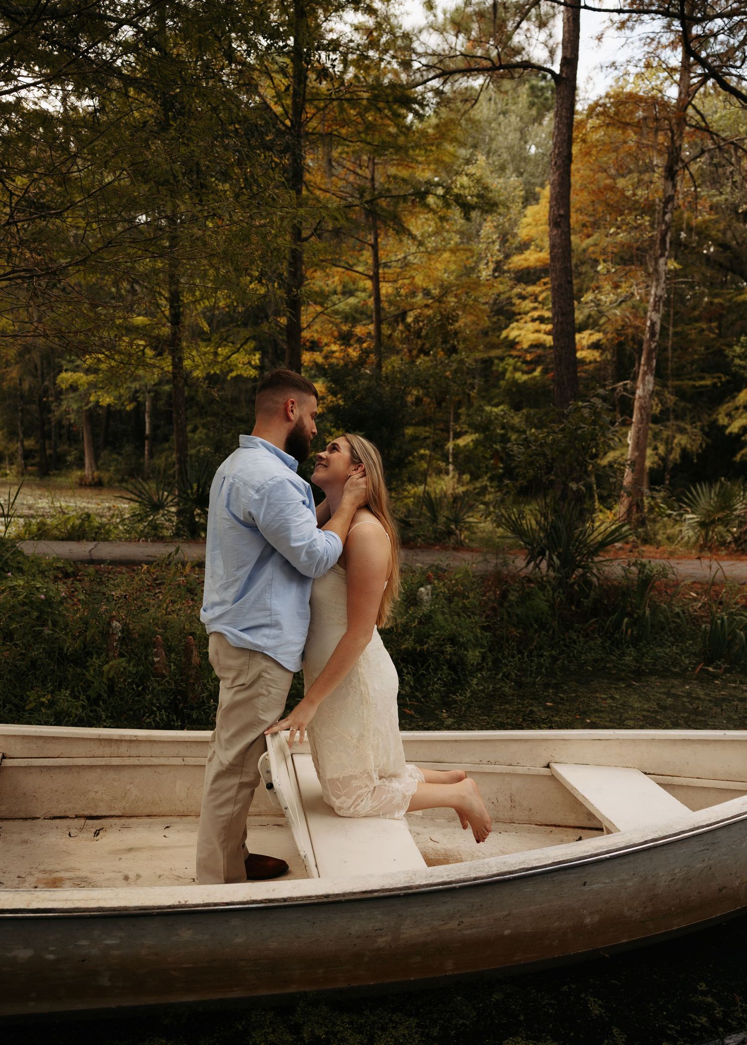 Couple sharing a romantic moment standing in a white rowboat surrounded by autumn trees near a tranquil lake.