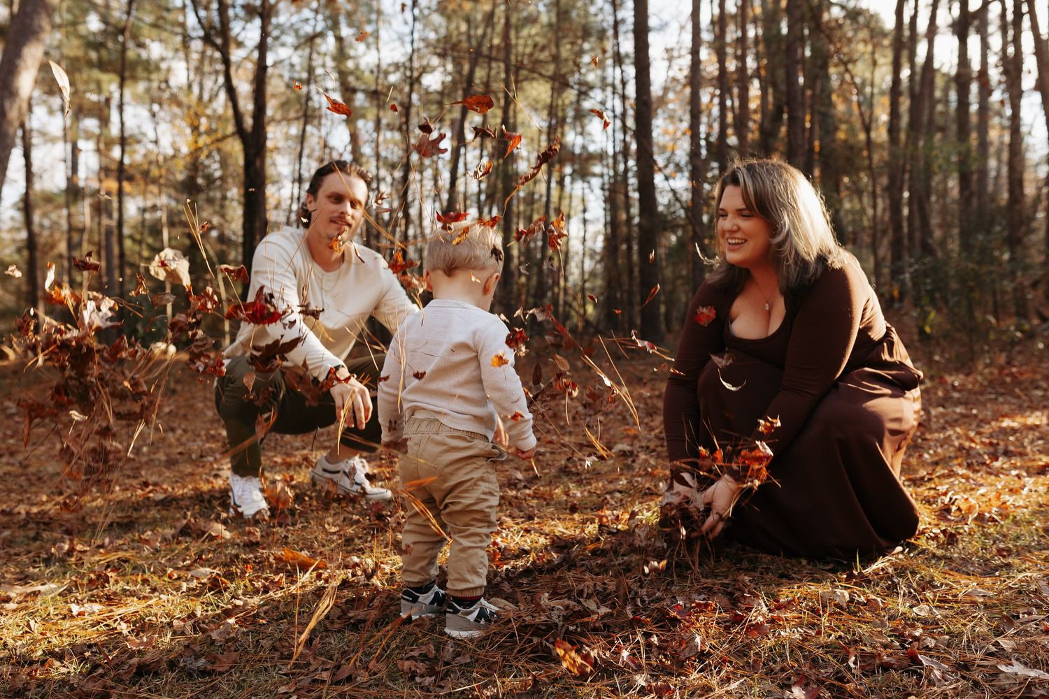Family playing with fall leaves in a sunlit forest, toddler reaching out to parents during autumn outdoor photo session.