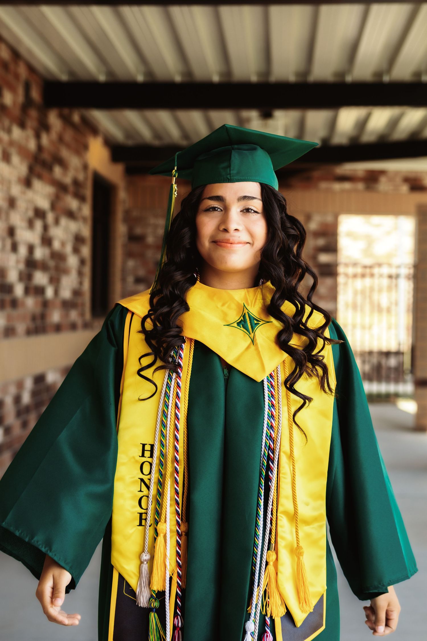 A graduate poses in green and gold honors regalia with curled hair and bright smile.