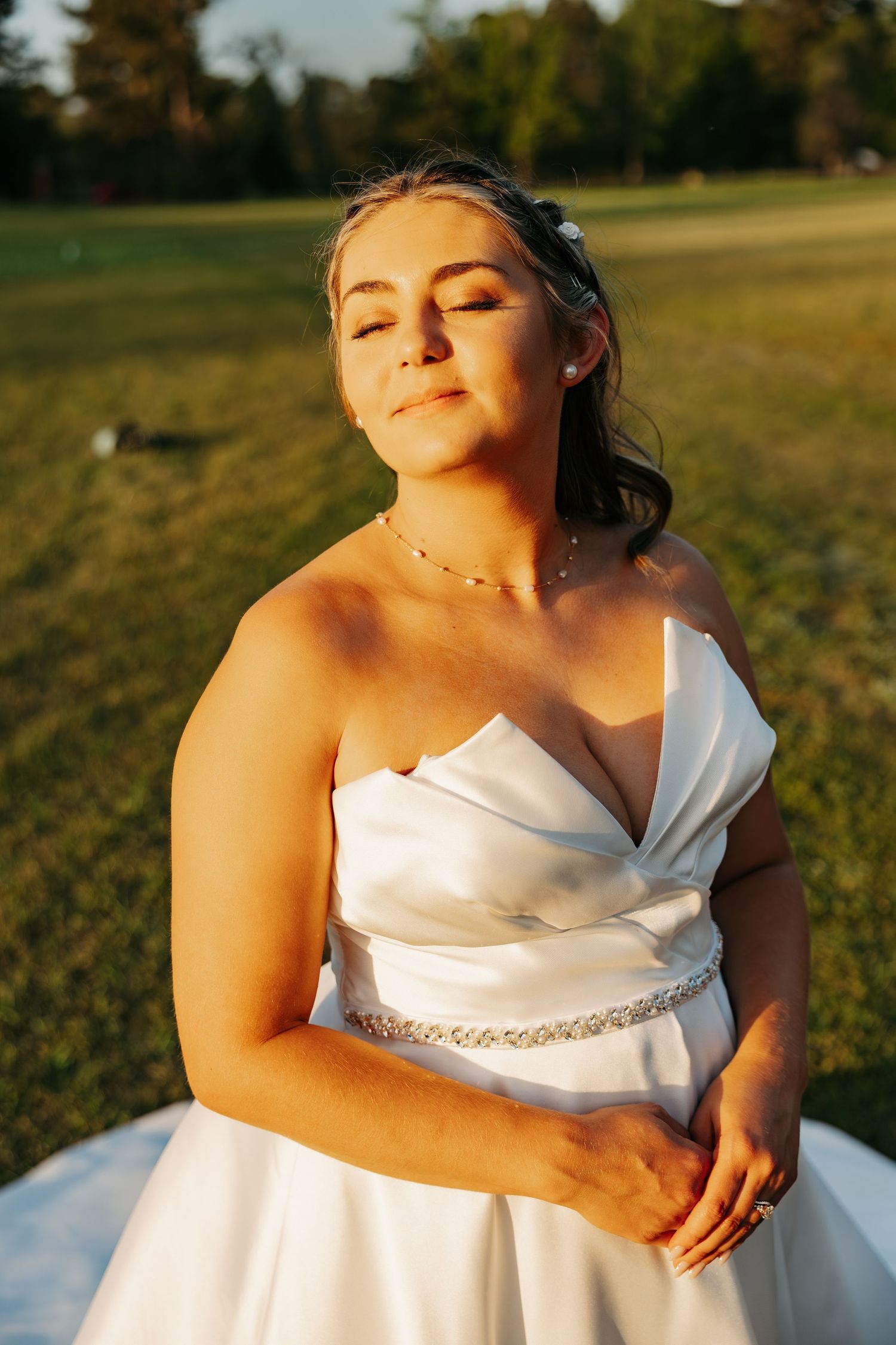 Radiant bride in strapless white gown with crystal belt basking in golden sunset light on green lawn.