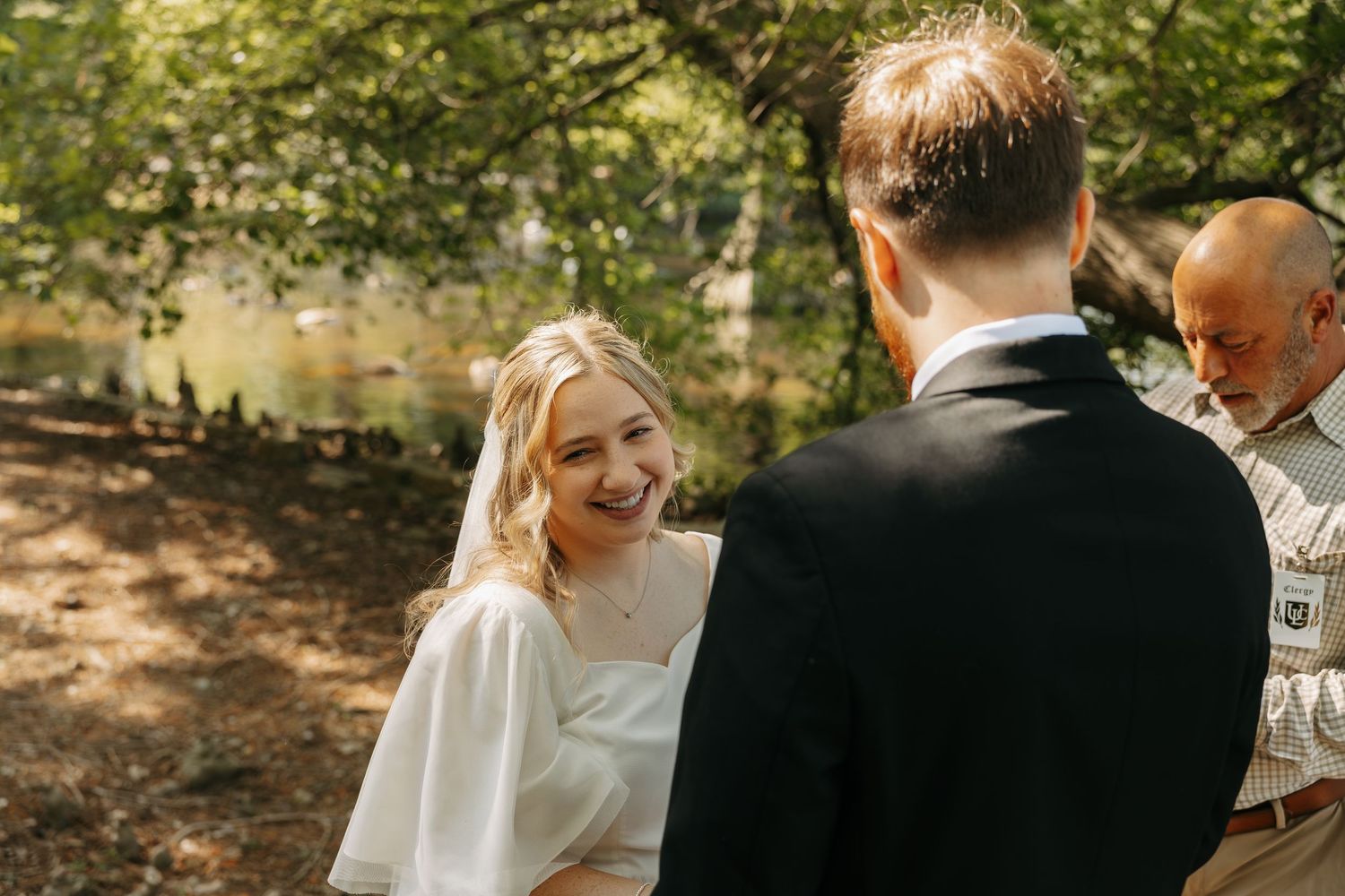 Smiling bride in white dress and veil exchanging vows with groom outdoors during intimate woodland wedding ceremony.