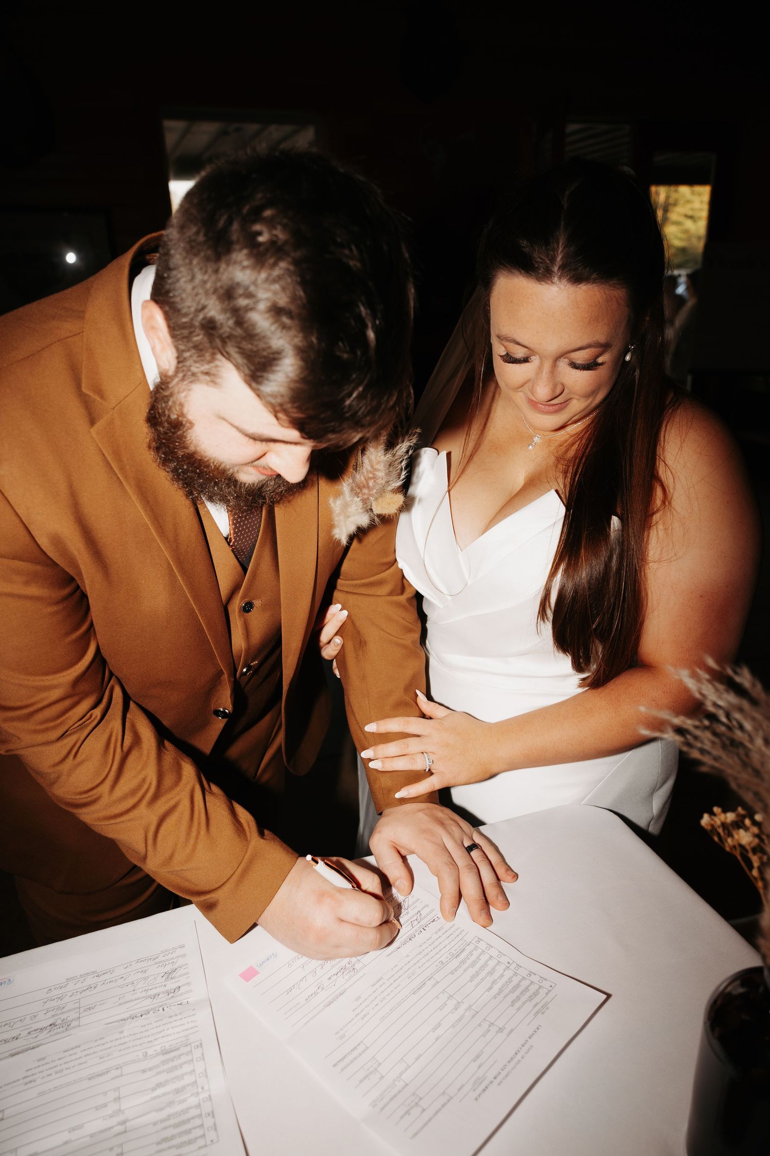 Bride and groom signing wedding certificate at reception table, groom in brown suit, bride in white dress.