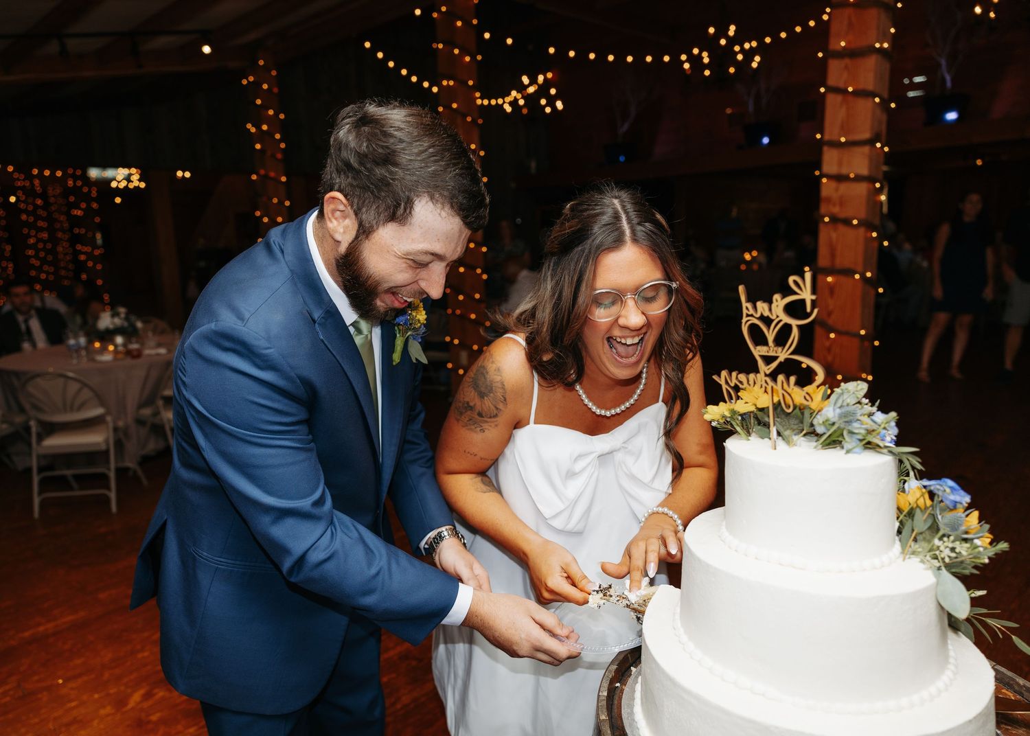 Laughing bride and groom cut white wedding cake together at indoor reception with string lights.