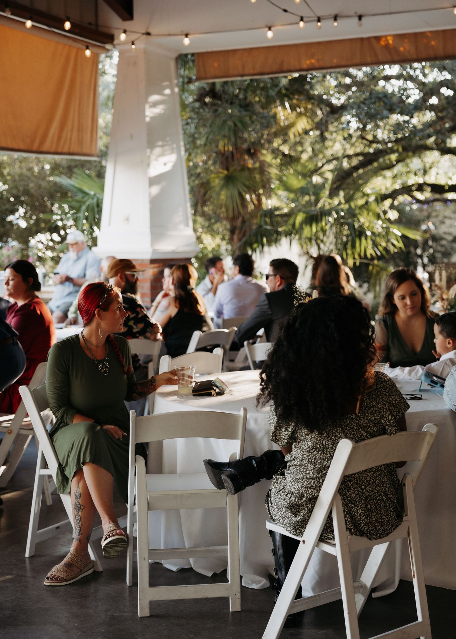 Wedding reception guests seated at round tables with white chairs under an open-air pavilion surrounded by trees.