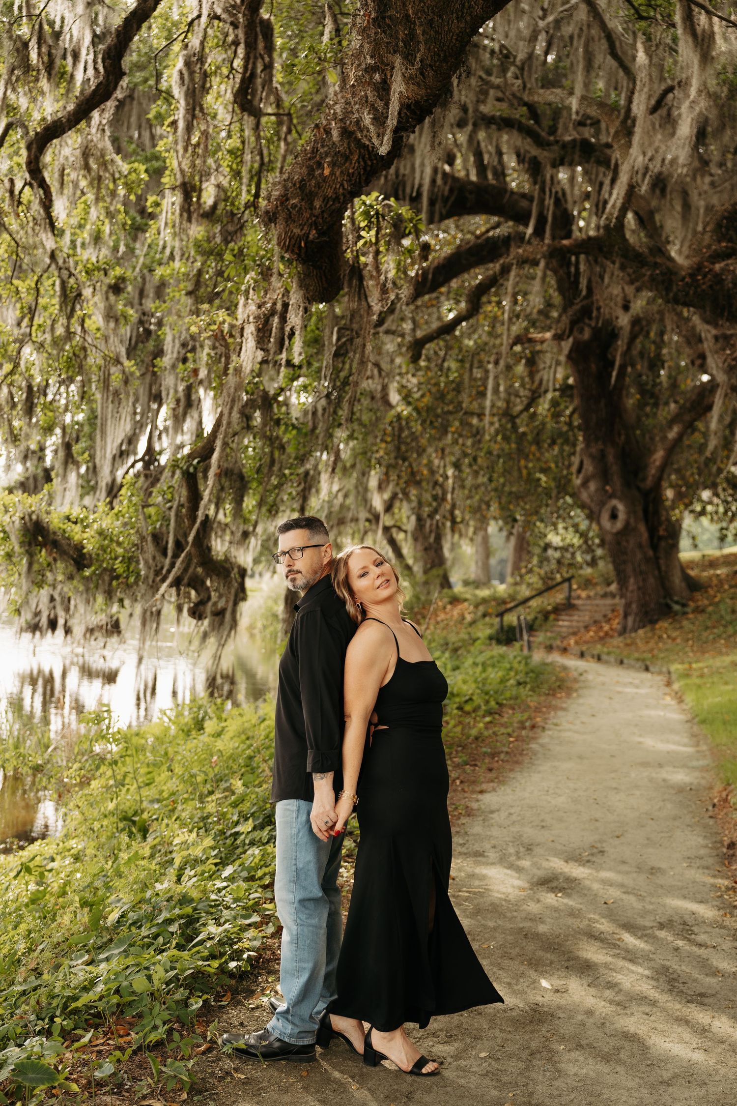 Couple in black outfits posing under moss-draped oak trees by a serene pond on a scenic garden path.