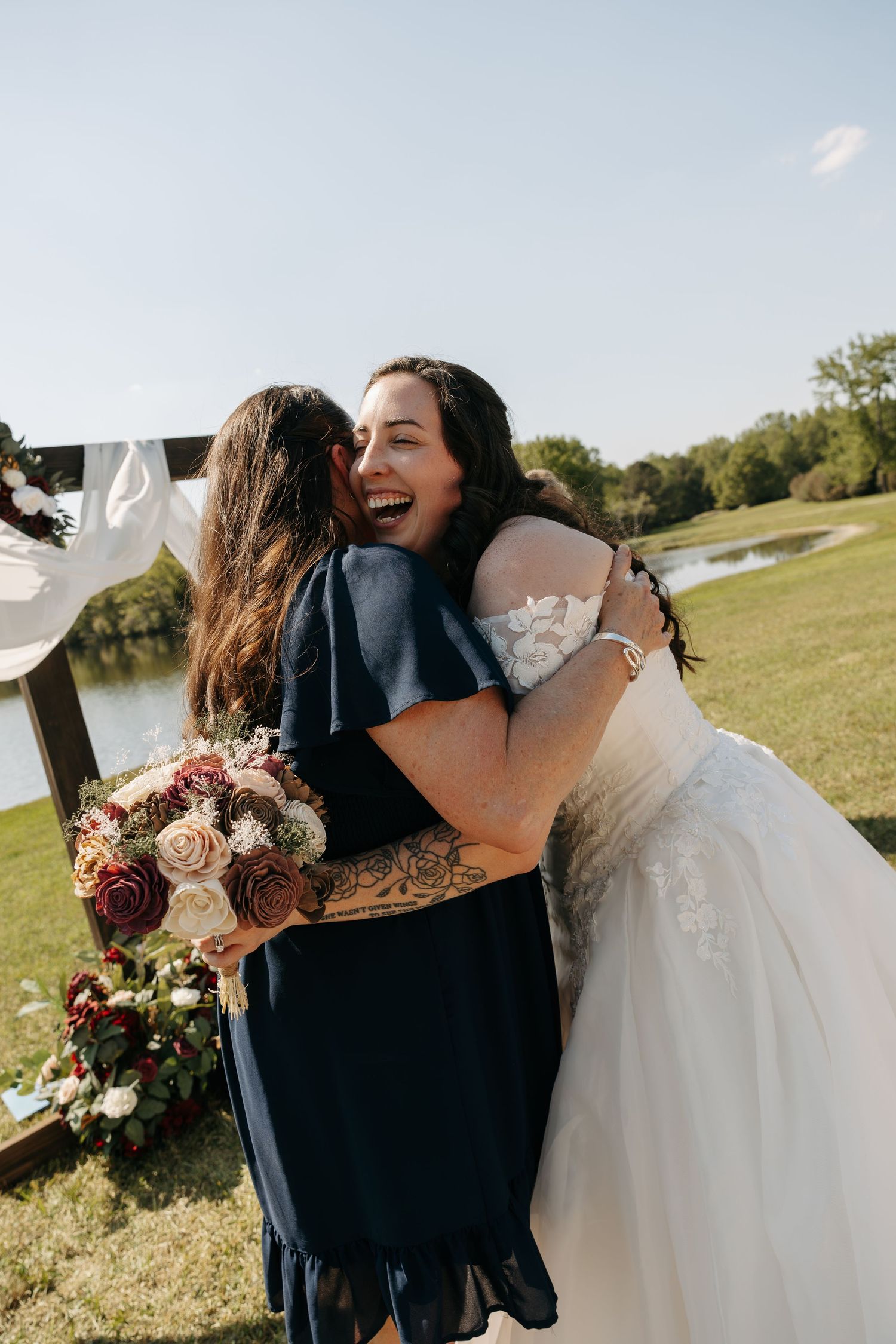 Bride in white gown laughing while hugging bridesmaid holding burgundy and pink bouquet outdoors.